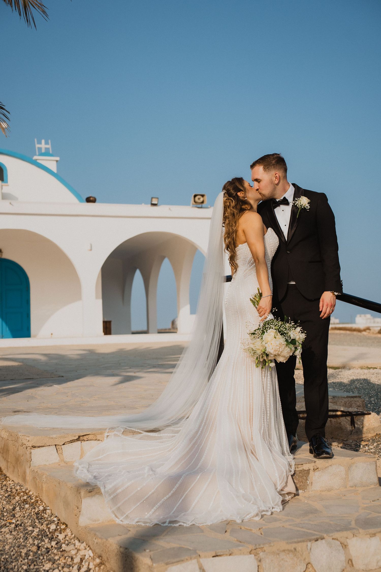 Bride and groom share a kiss outside a white Mediterranean chapel with blue doors on their wedding day.