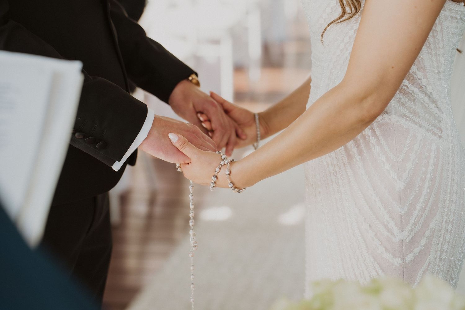 Bride and groom holding hands with rosary beads during outdoor wedding ceremony exchange.