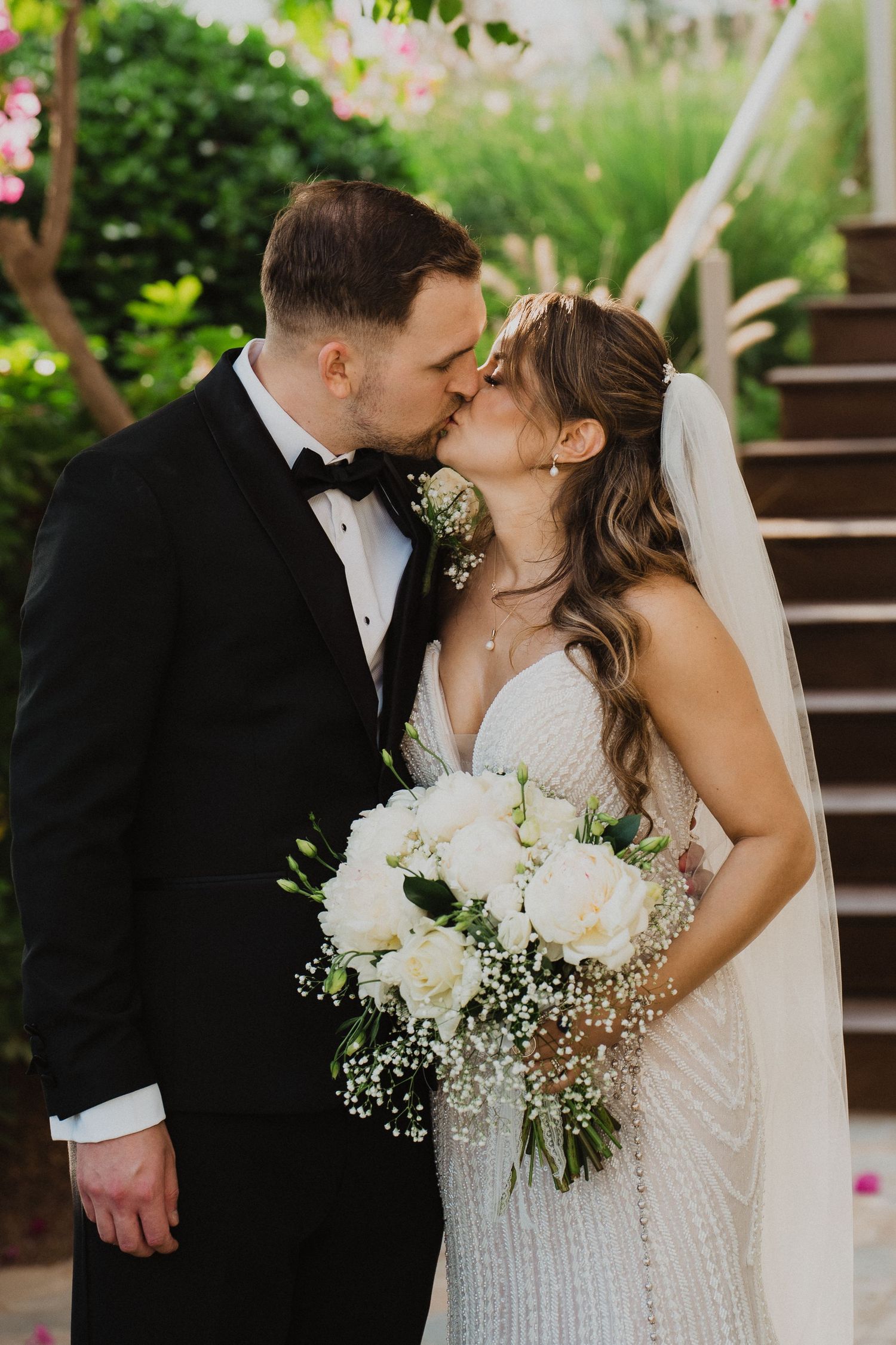 Bride and groom share romantic kiss as bride holds white rose and baby's breath bouquet outdoors.