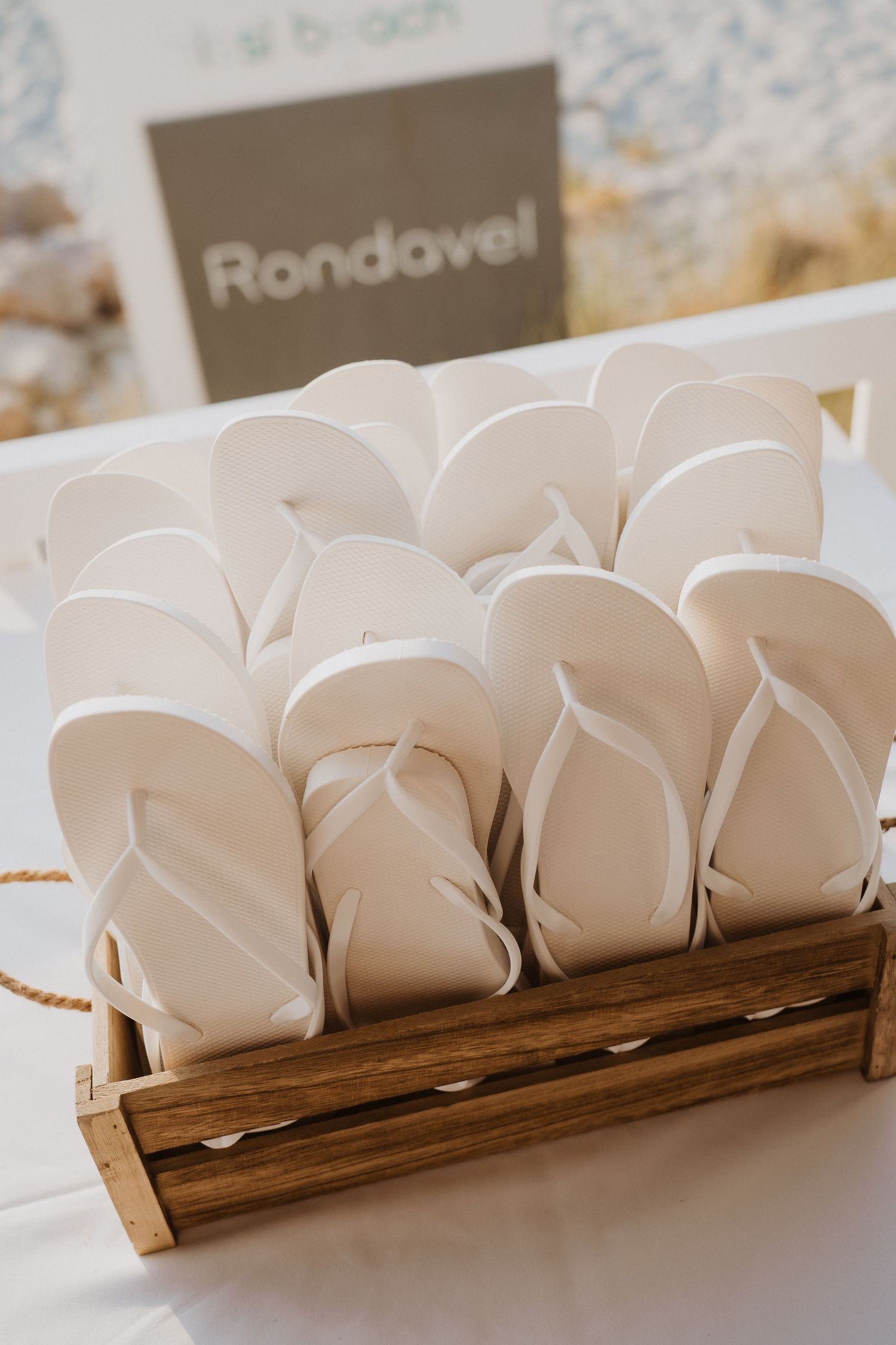 White flip flops displayed in a wooden crate labeled Rondavel as wedding guest favors at a beach venue.
