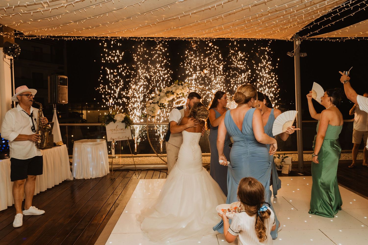 Wedding reception dance floor with sparklers, bride in white gown, bridesmaids in blue and green dresses, festive celebration.