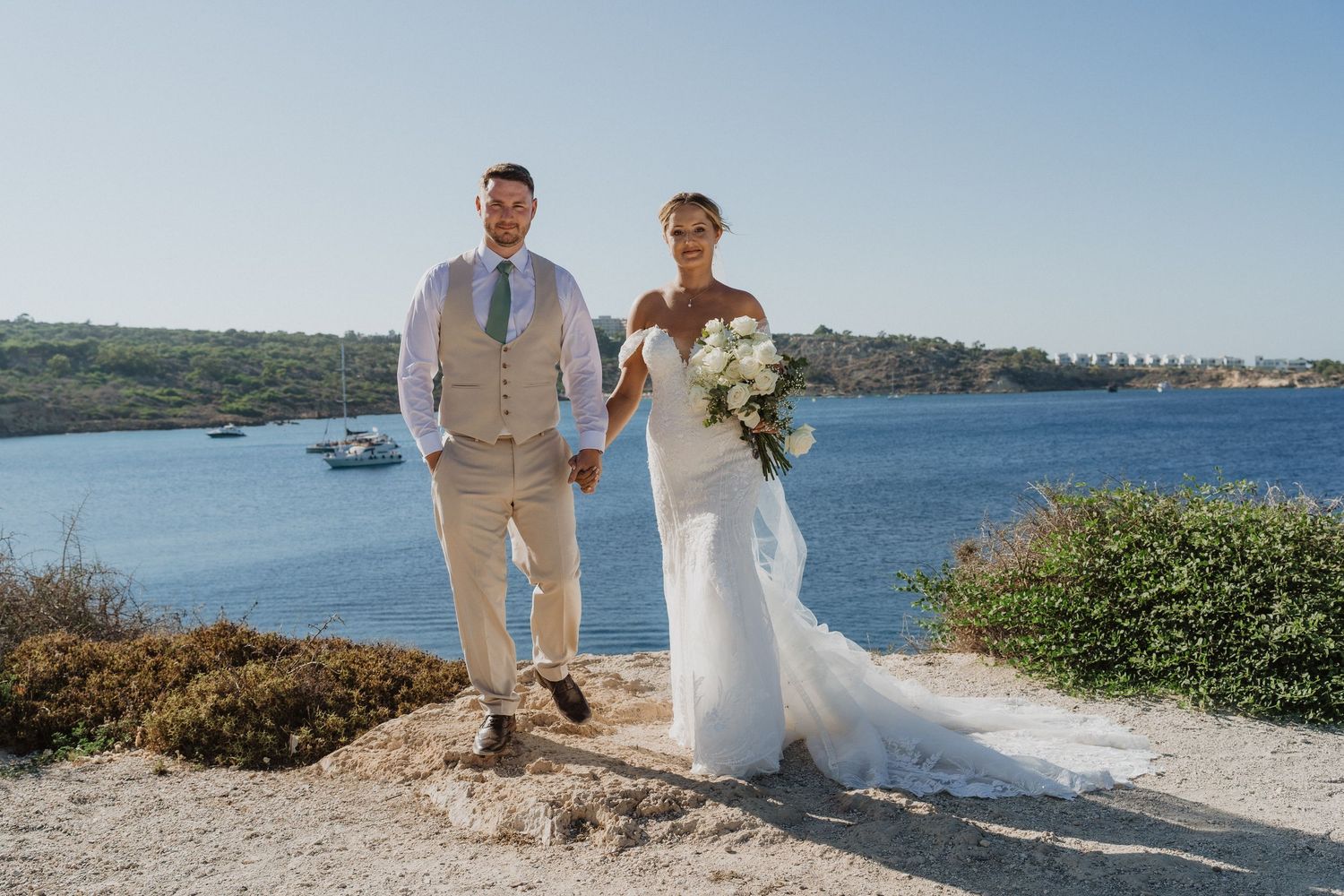 A couple in wedding attire stands together on a scenic coastal cliff overlooking a blue bay on a sunny day.