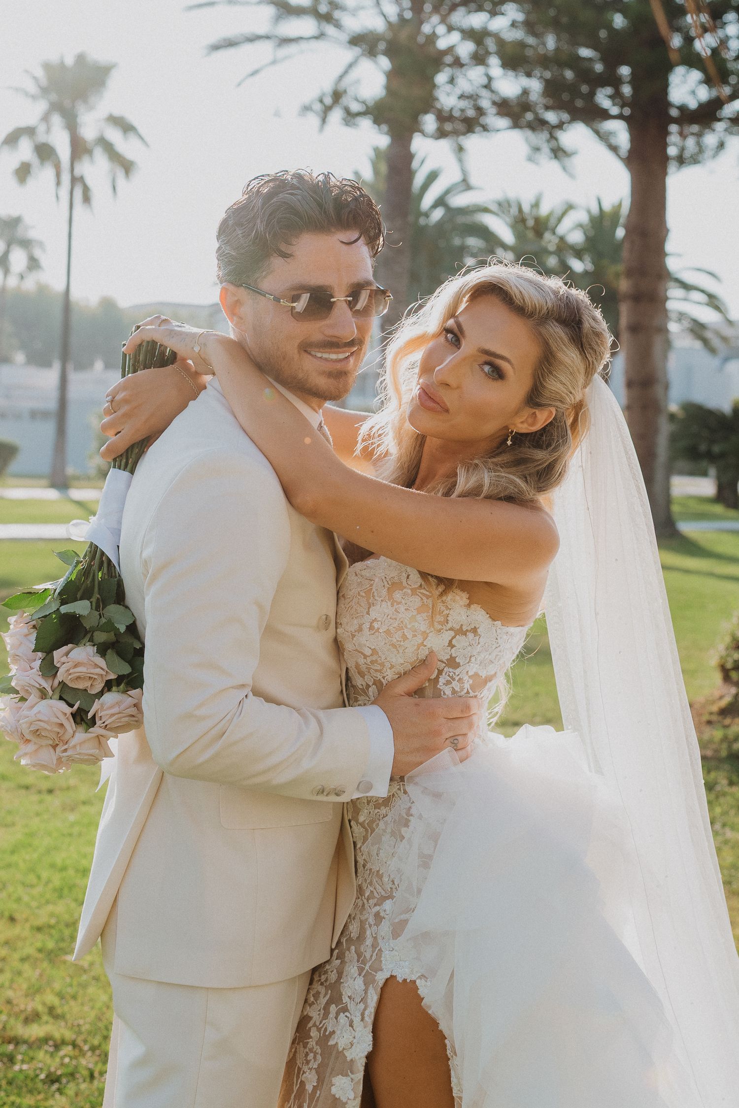 A couple shares romantic moments in white wedding attire on a sunny day with palm trees in the background.