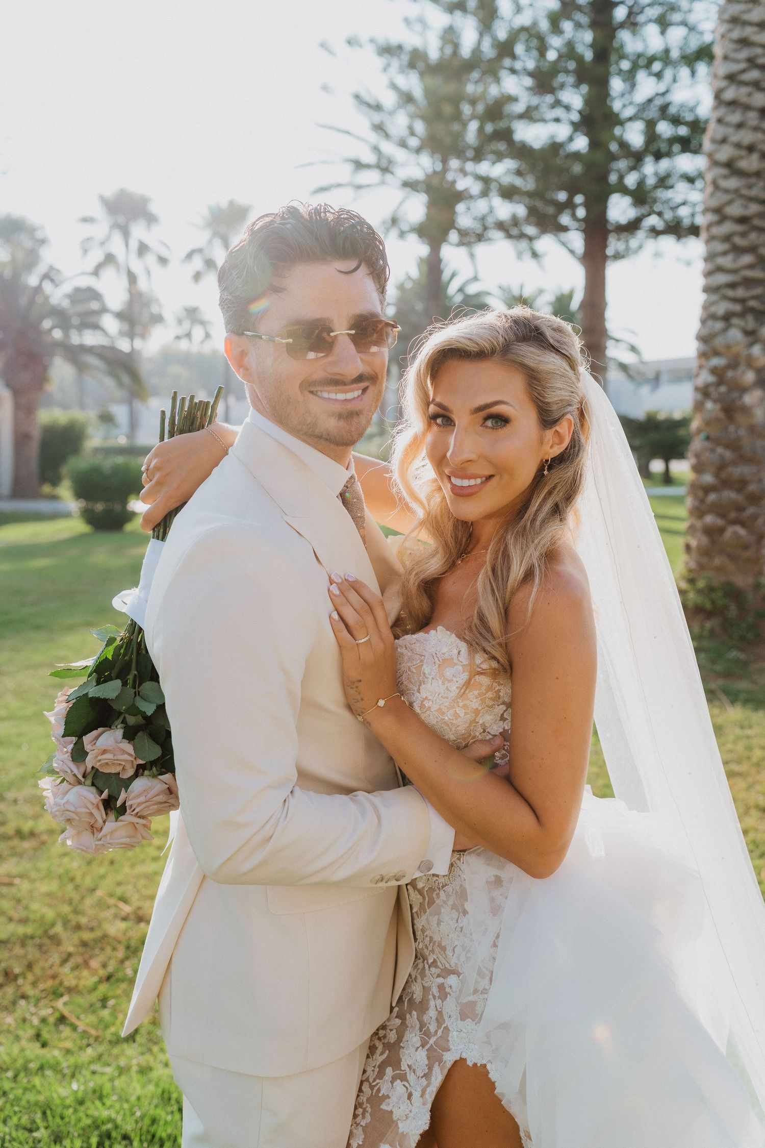 A newlywed couple poses outdoors on their wedding day in a sunny garden setting with palm trees.