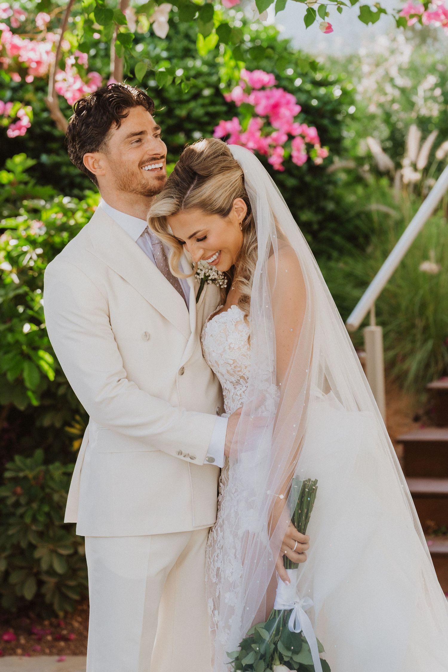 A romantic wedding couple embraces in a garden setting with pink bougainvillea flowers blooming in the background.