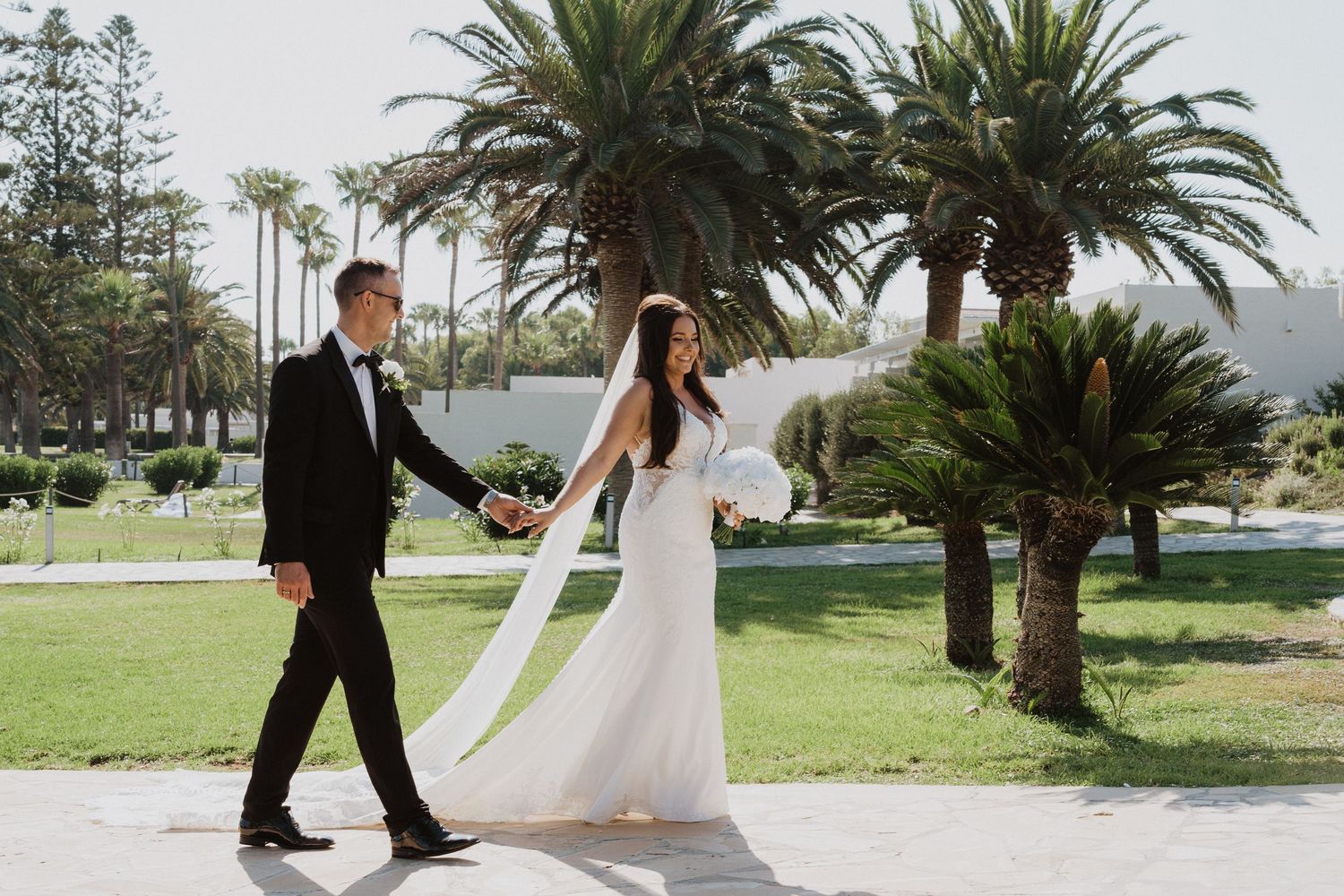 A couple strolls through a lush garden setting with palm trees during their wedding day.