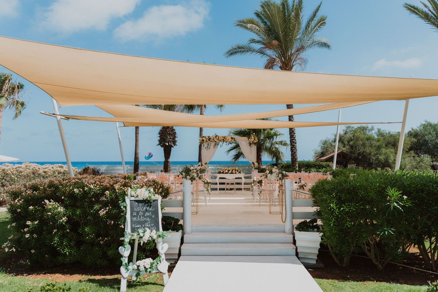 Outdoor wedding ceremony setup with beige canopy and palm trees at a tropical beach venue under blue skies.