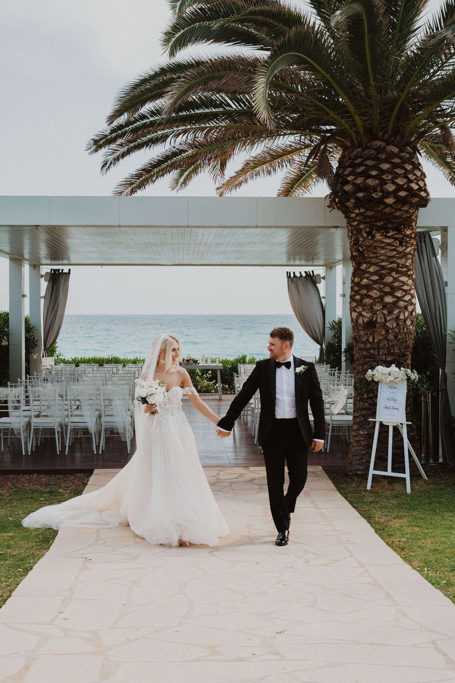 Newlyweds walk hand in hand along a stone path with palm trees and ocean views.