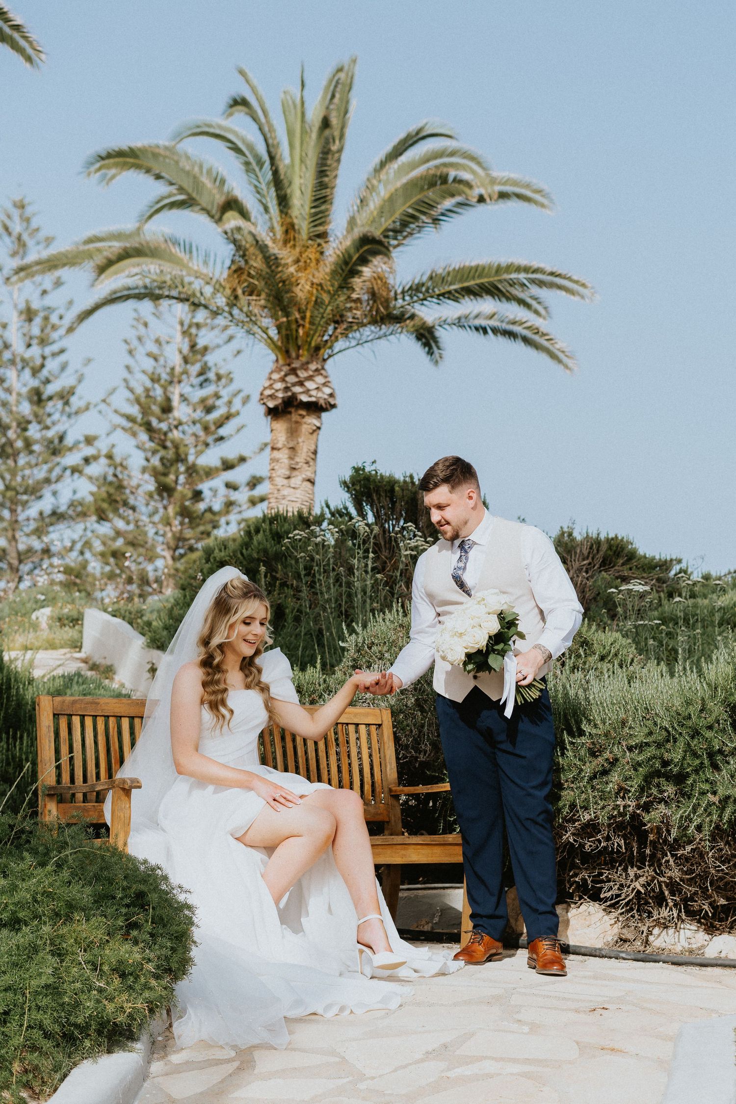Newlyweds share romantic moment on garden bench beneath towering palm tree in tropical wedding setting.