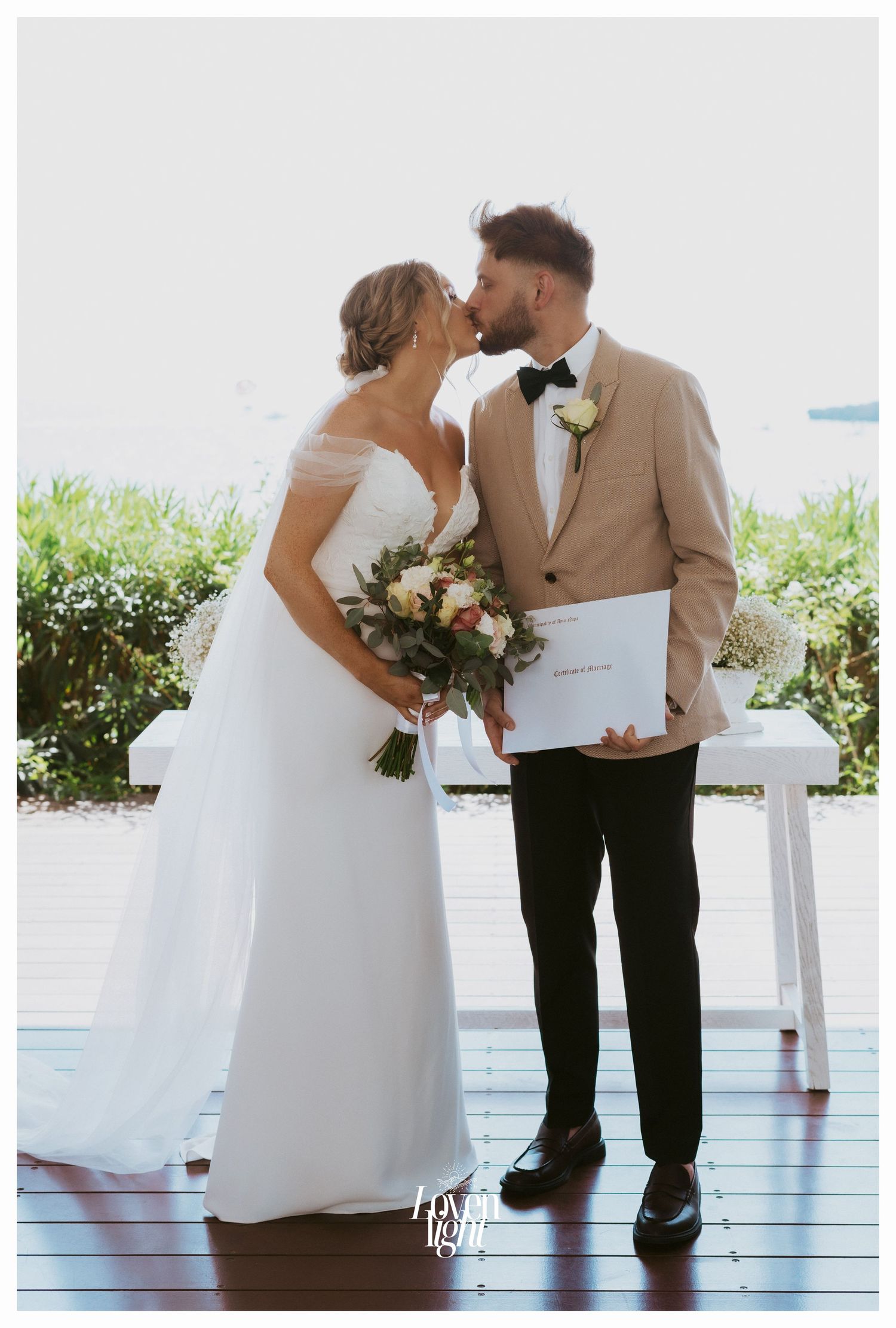Elegant couple with wedding bouquet signing documents on white table outdoors.