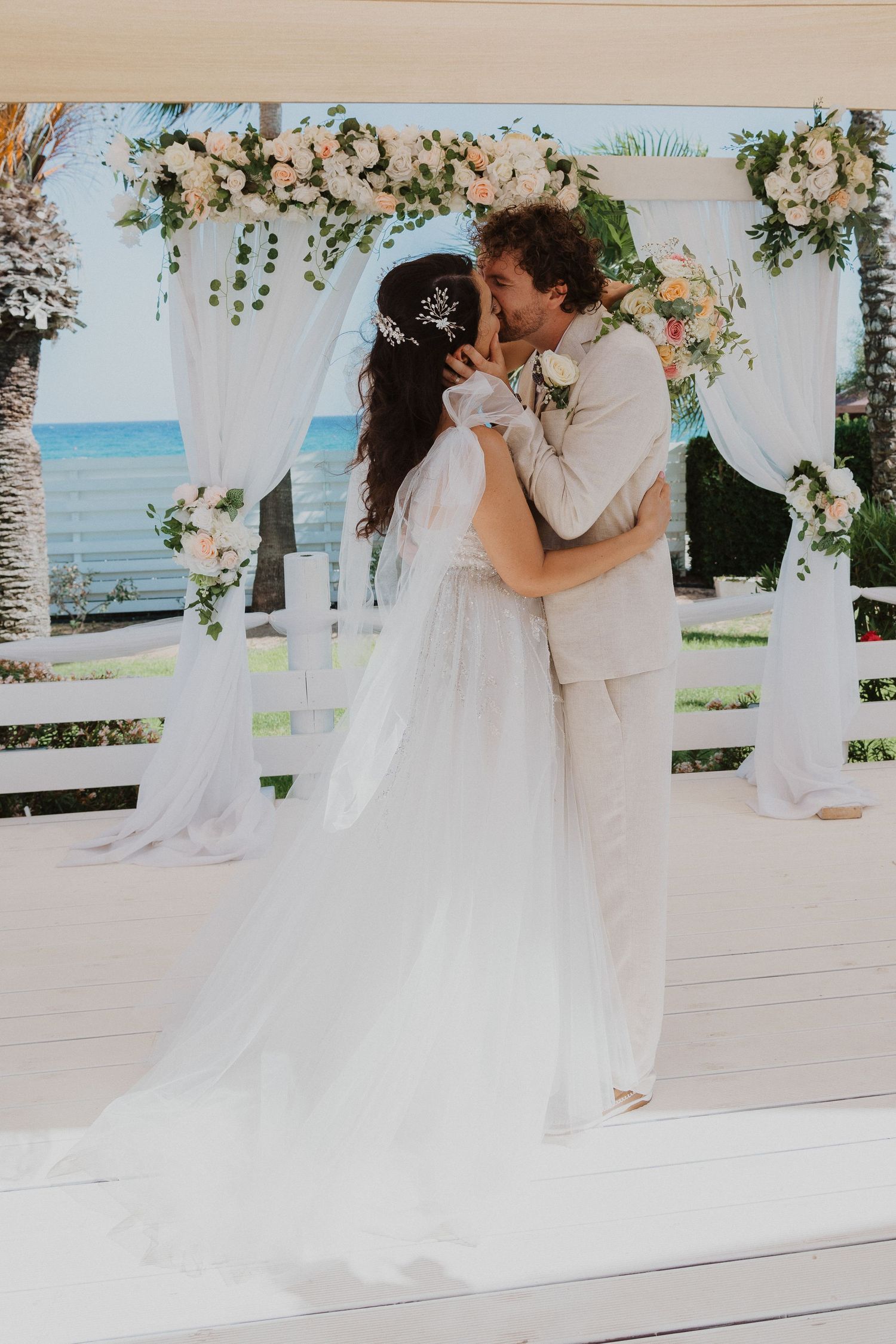 Romantic wedding portrait under floral arch with white drapes and beachfront view.
