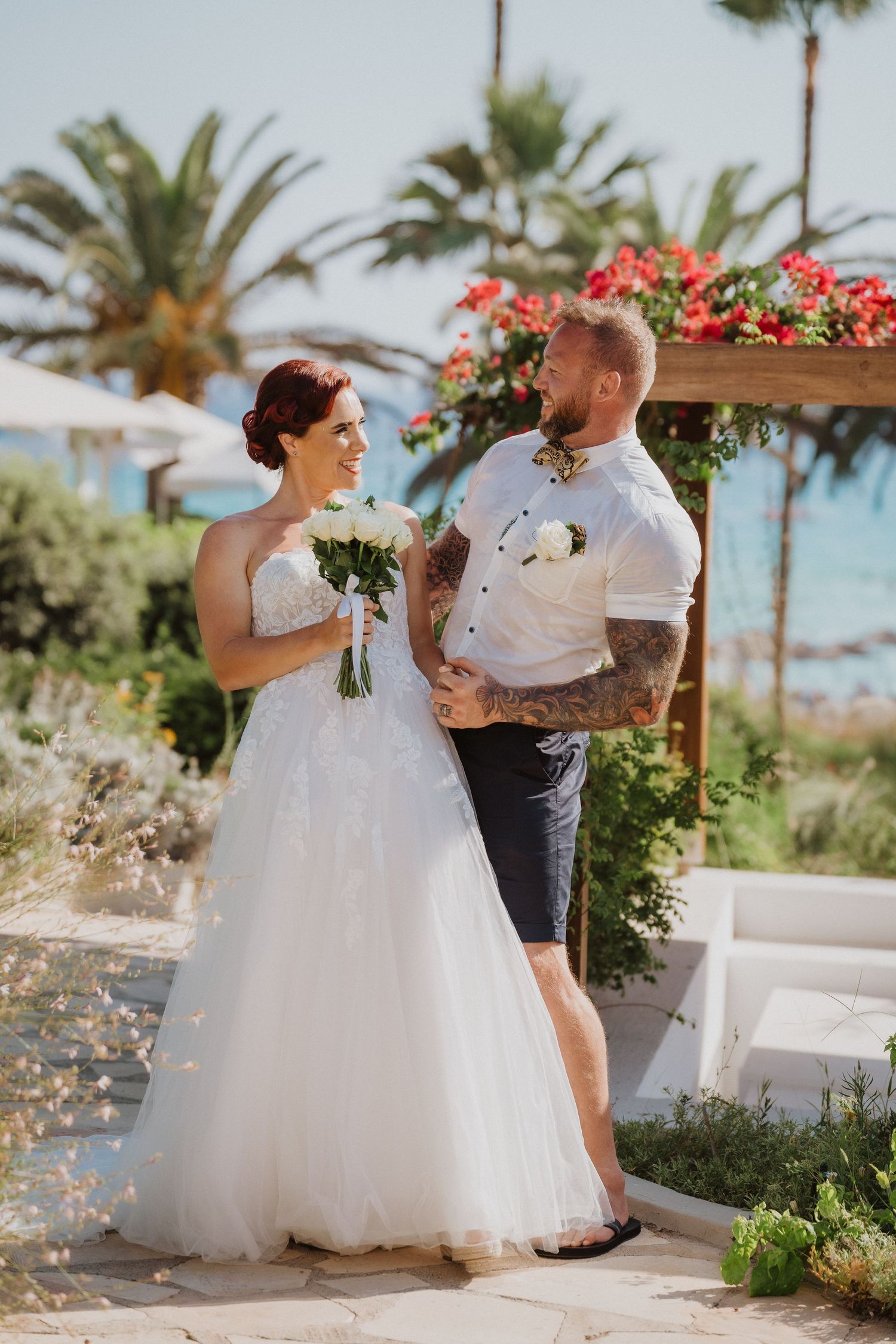 Wedding couple stands together under wooden arch decorated with red flowers in tropical setting.