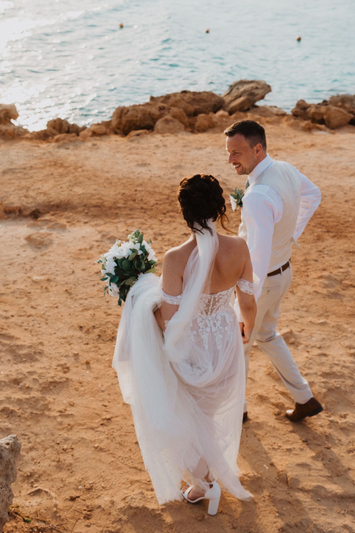 A couple in wedding attire stands on a coastal cliff with ocean views and rocks during sunset.