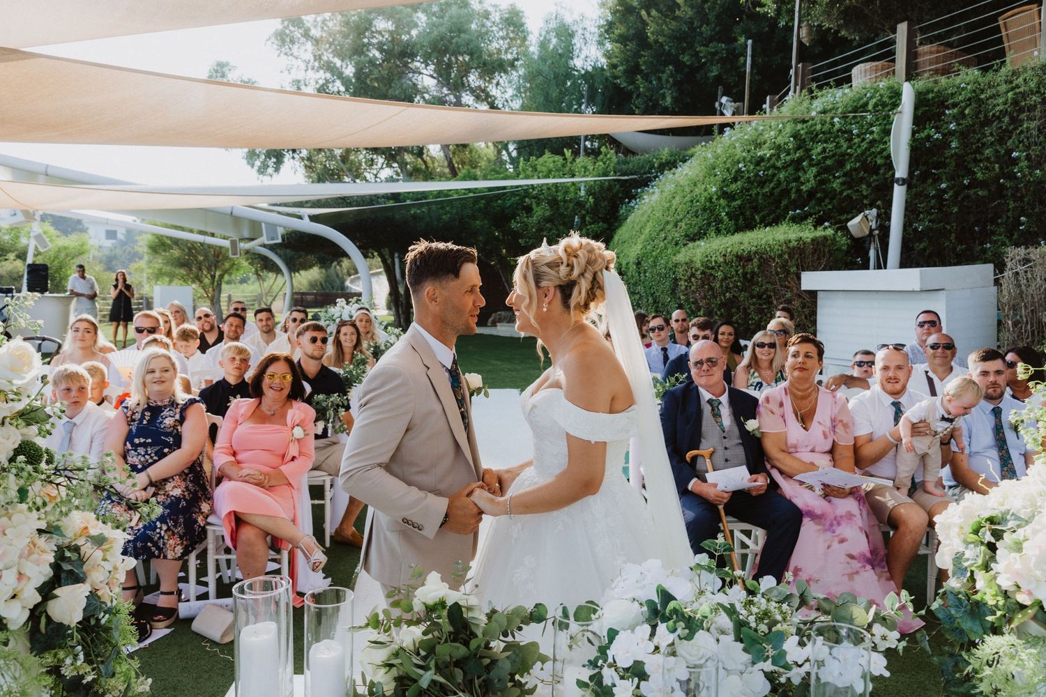 Romantic outdoor garden wedding ceremony with elegant floral arrangements and guests in pink and white attire.