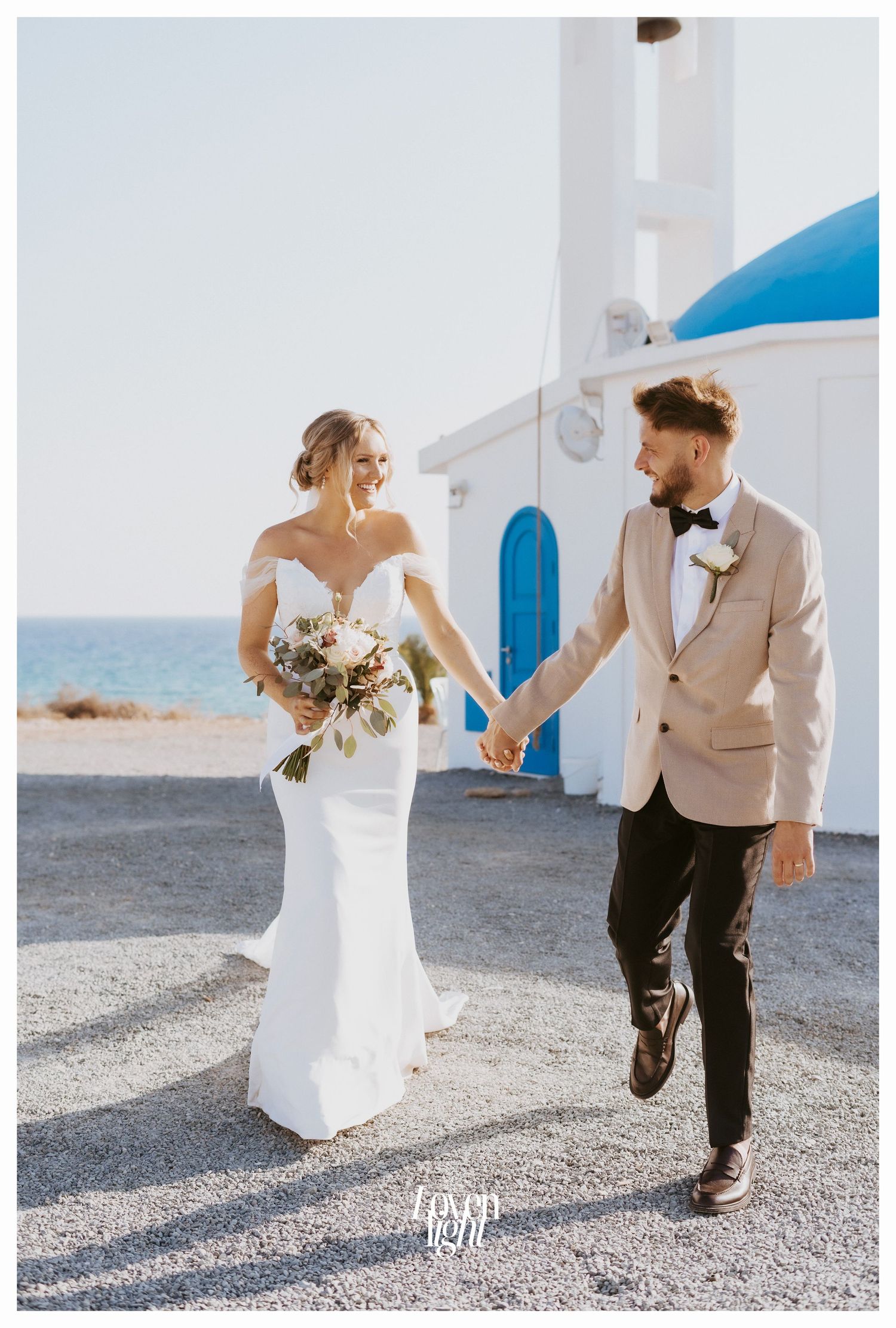 Joyful wedding couple holding hands while walking near coastal Mediterranean chapel.