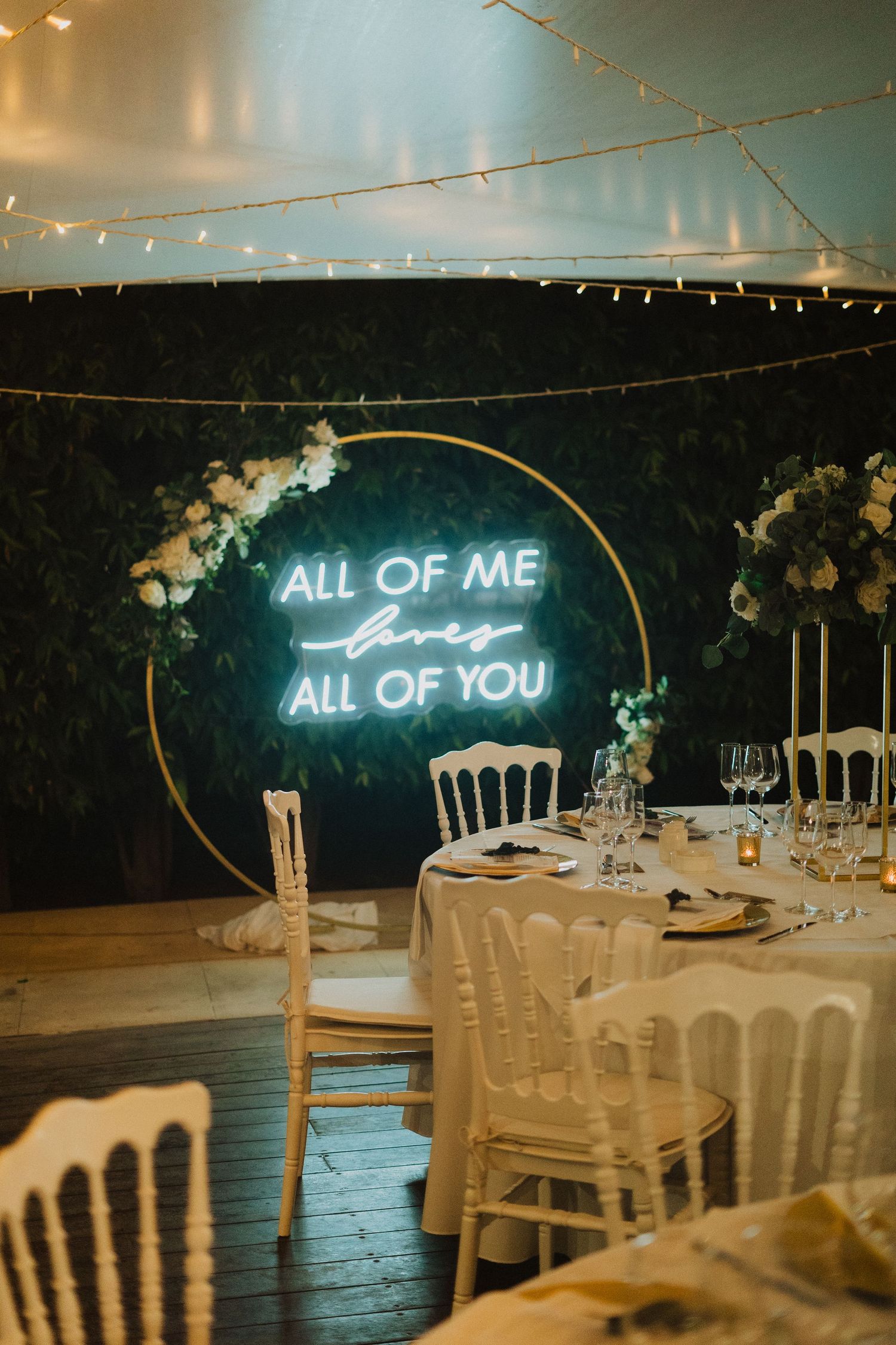 Romantic wedding reception setup with neon sign reading 'All of me All of you' above elegant white table settings.