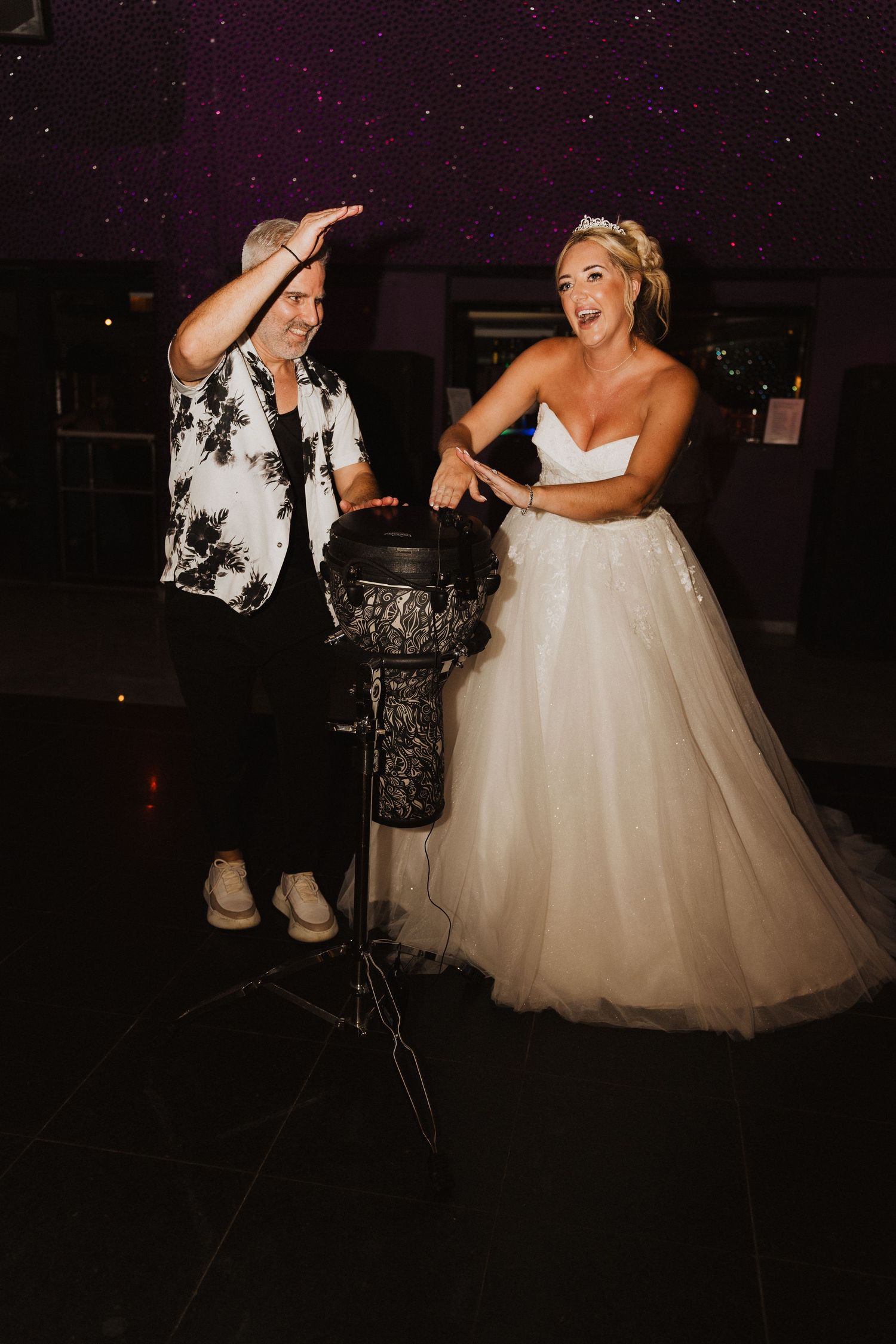 A bride in a white ball gown shares a joyful dance moment with a guest in a floral shirt at a wedding reception.