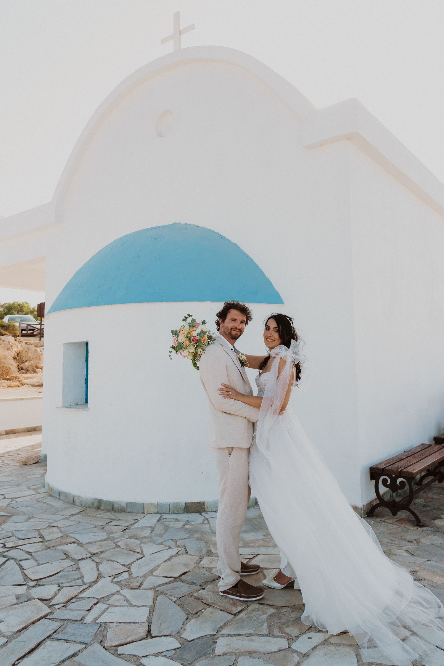 Wedding couple poses in front of a traditional Greek church with white walls and blue dome.