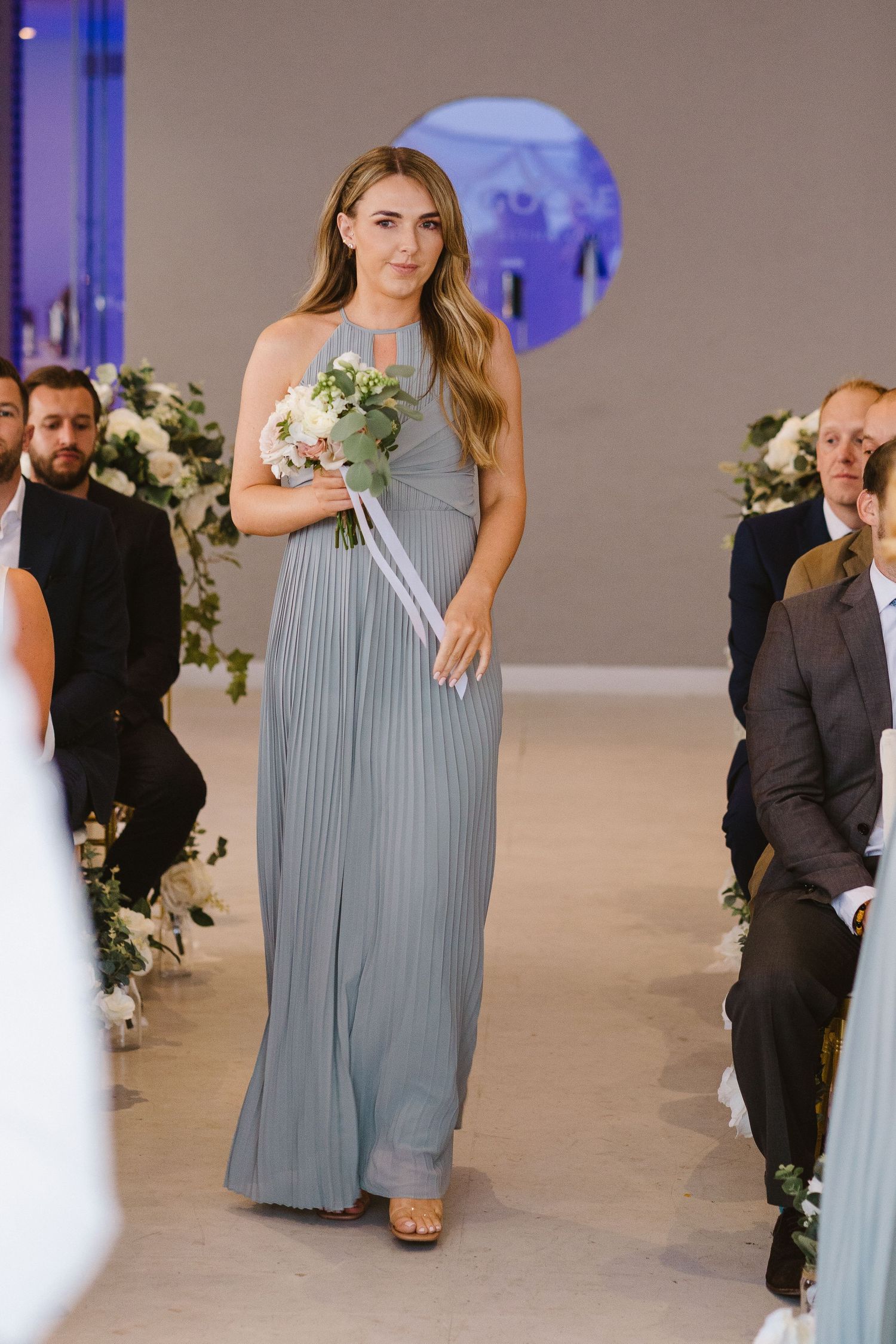 Bridesmaid in a flowing grey gown walks down the aisle carrying a small white bouquet.