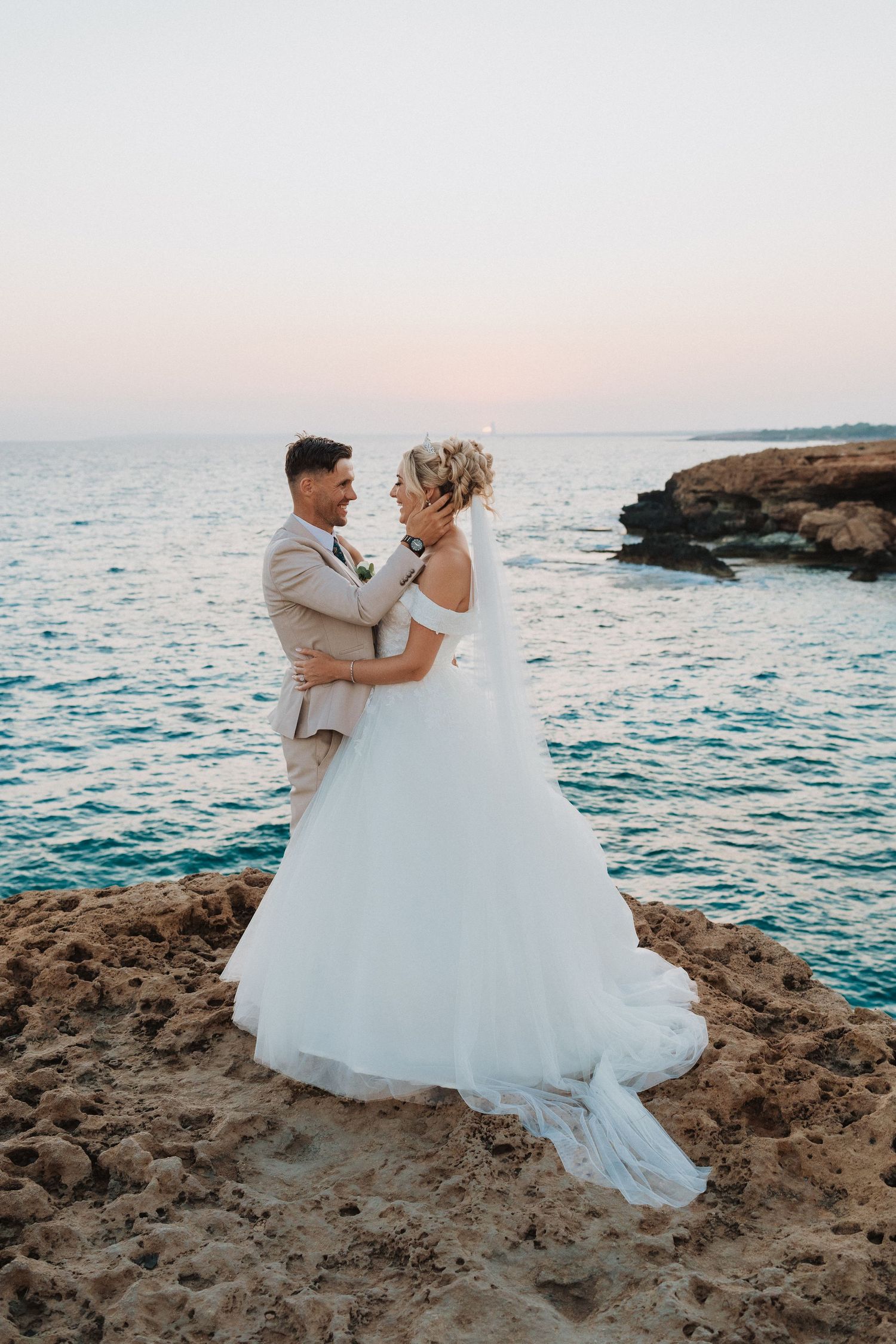 A romantic wedding photo of a couple embracing on coastal rocks at sunset with turquoise waters in the background.