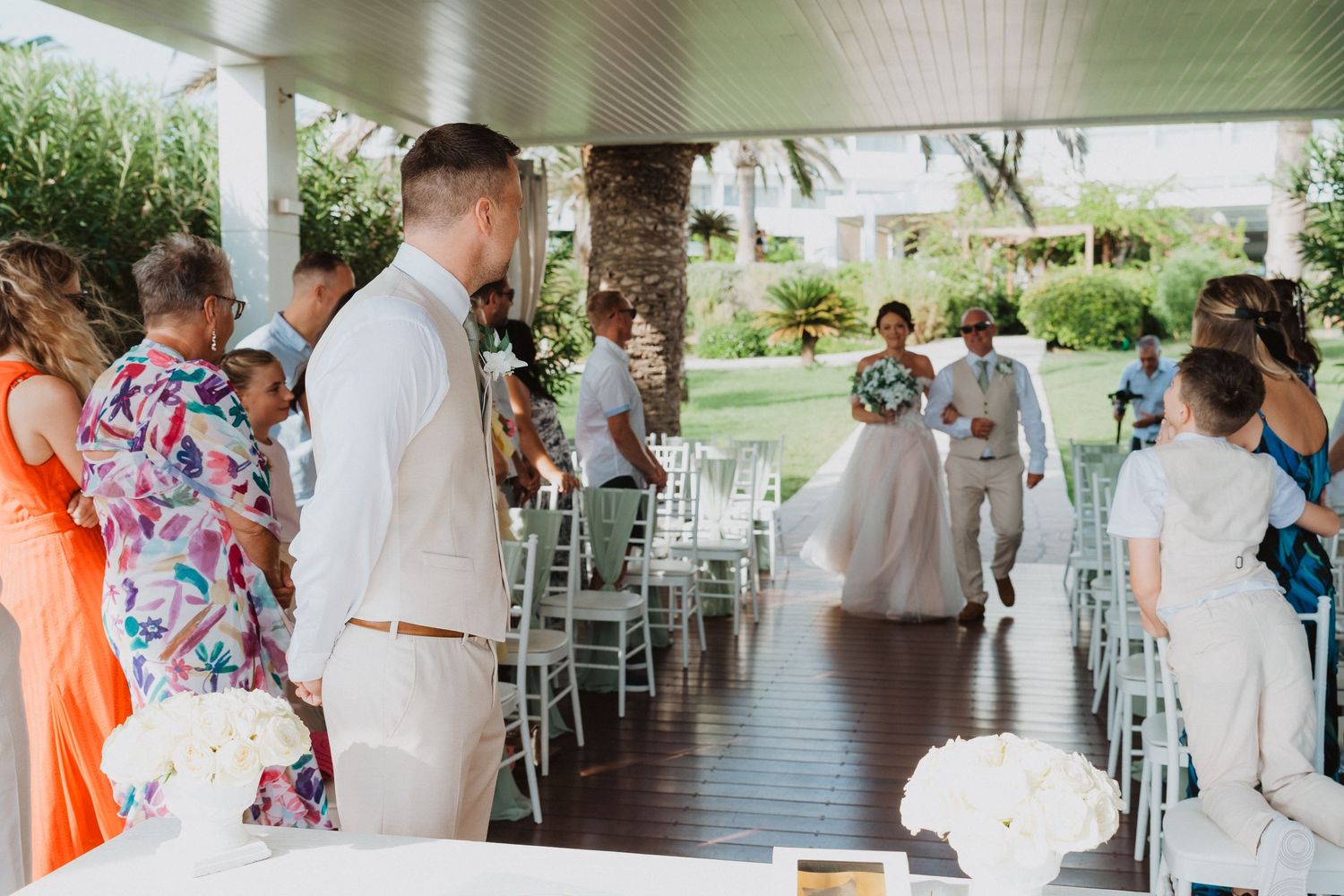 Wedding ceremony guests gather under a covered outdoor venue with white chairs and tropical landscaping.
