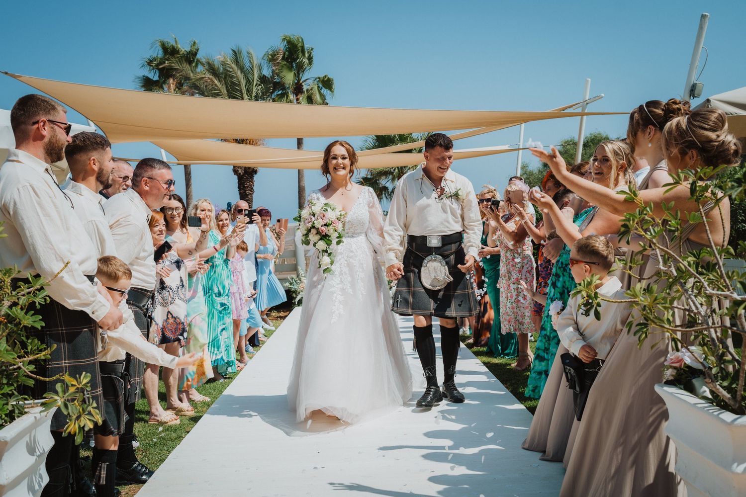 Wedding guests line outdoor aisle as couple walks between them during tropical beach ceremony.