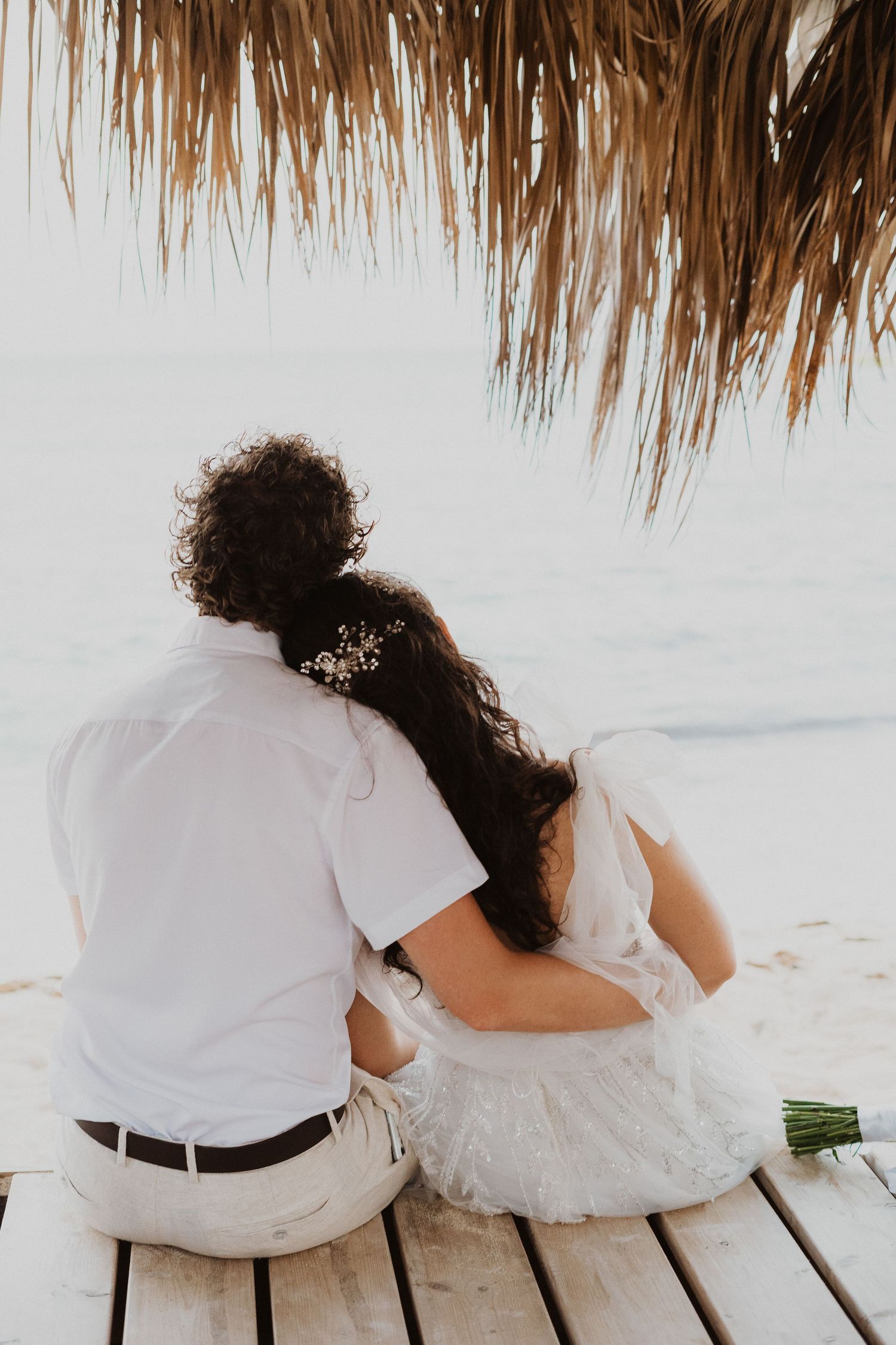 A couple shares an intimate moment on a wooden deck under a thatched palapa by the beach.