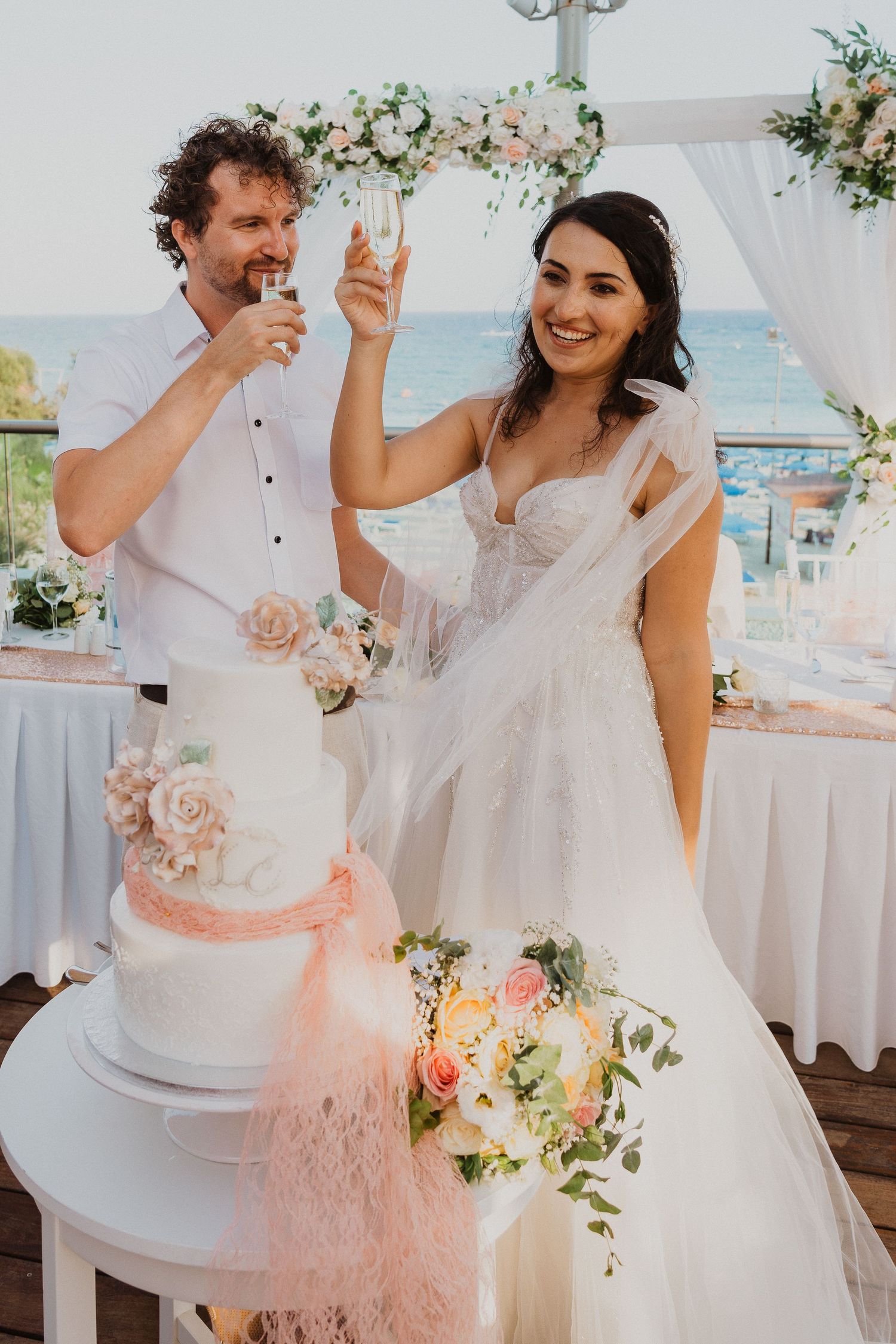 Newlyweds share a champagne toast beside their wedding cake at a seaside celebration.