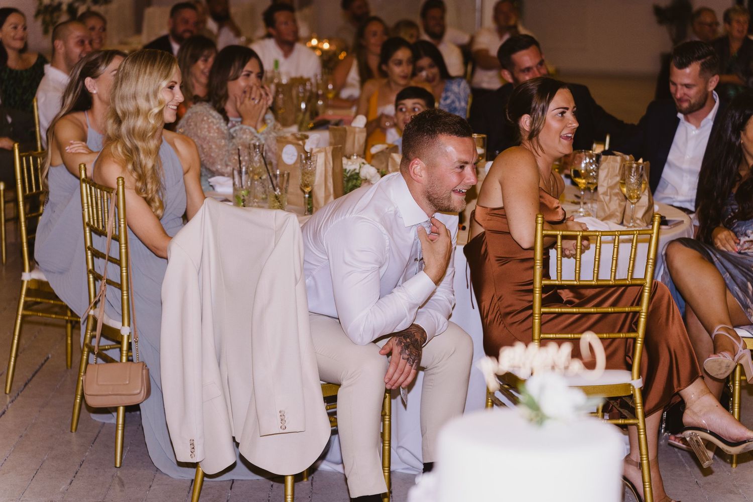Wedding guests enjoying speeches and celebration at elegant reception with gold chairs.