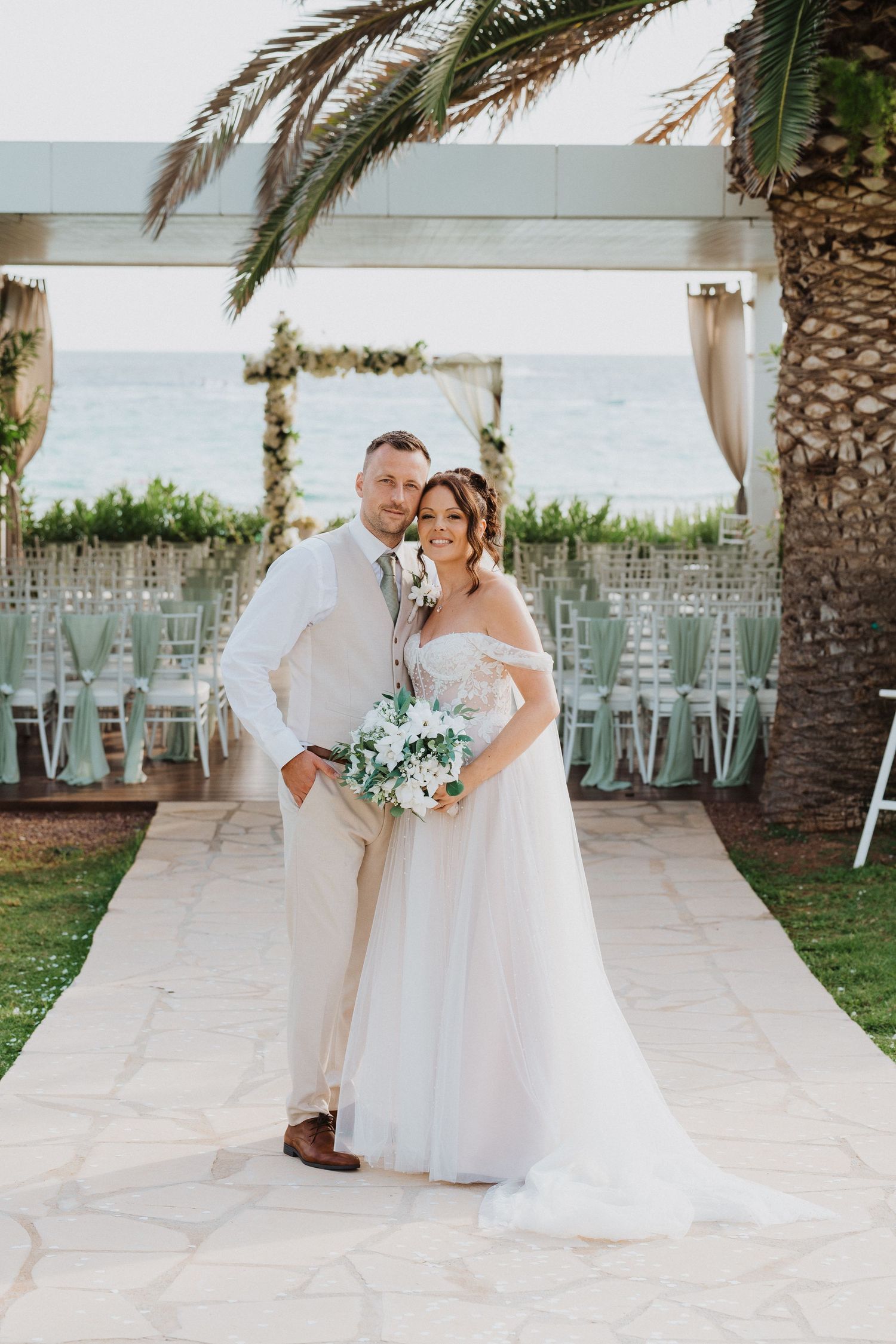 A white wedding ceremony setup with palm trees and ocean views featuring a decorated arch and mint green chairs.