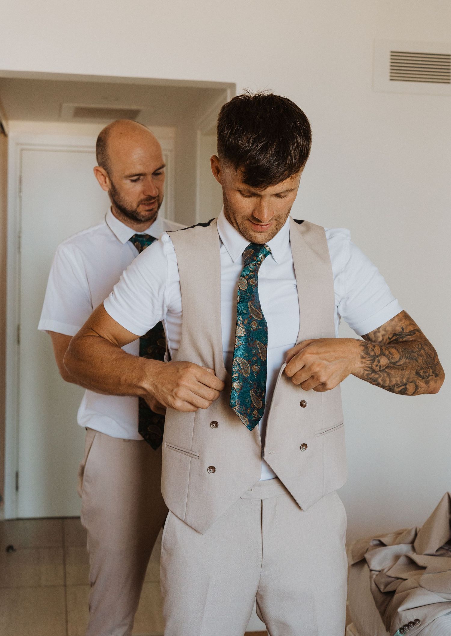 Two people getting dressed in formal attire with light colored suits and ties.