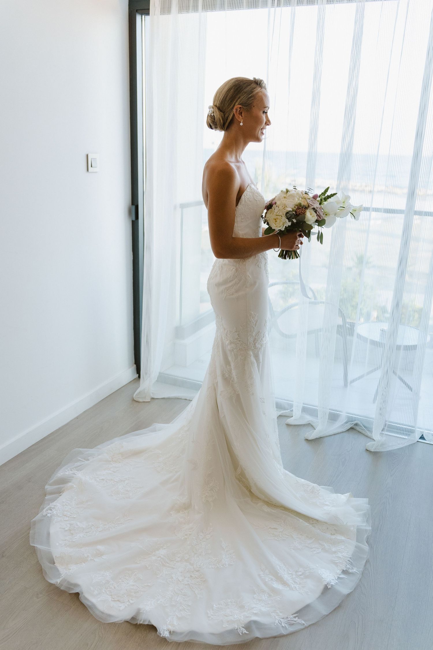 A bride in a fitted lace wedding gown holds a white bouquet while posing by a window.