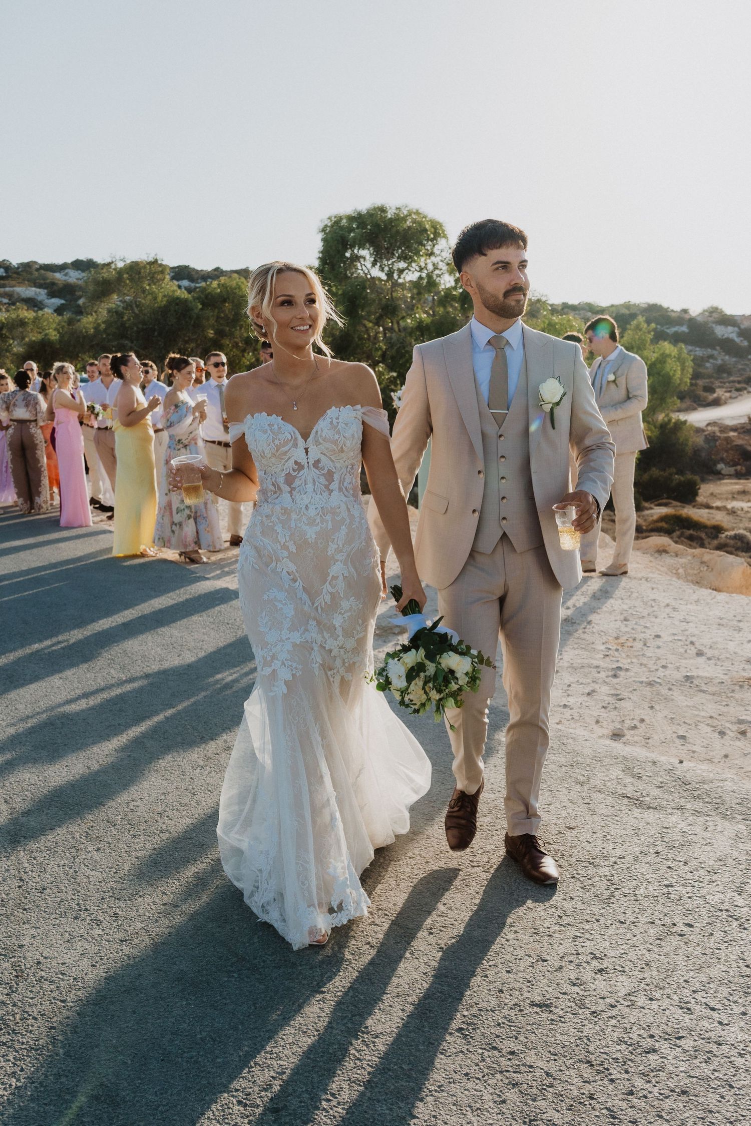 Wedding guests in summer attire follow the newlyweds along a scenic coastal pathway.