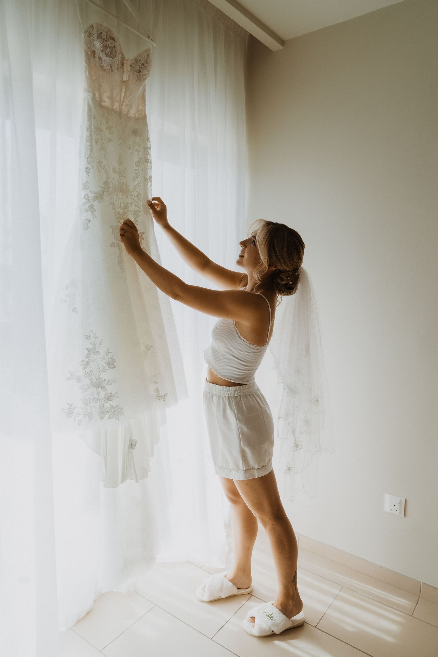 Person in white loungewear reaches up to touch wedding dress hanging by window.