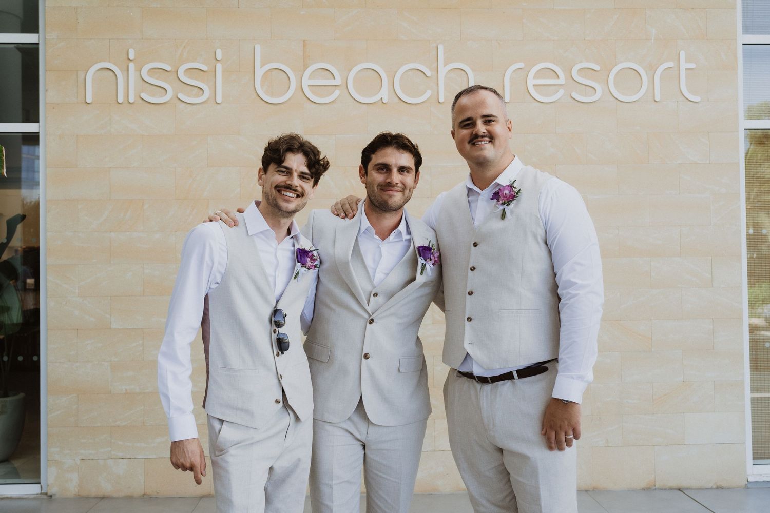 Three men in light colored suits pose together in front of the Nissi Beach Resort sign.