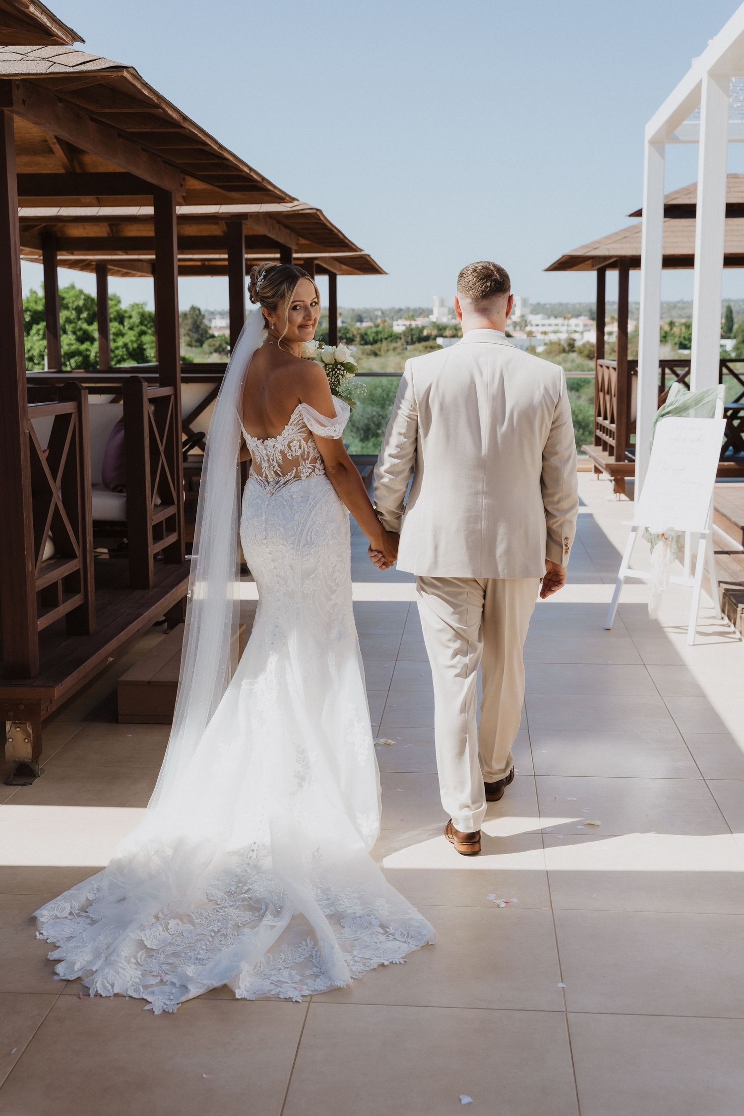 A couple in wedding attire walk along a covered terrace with scenic views of a tropical resort destination.