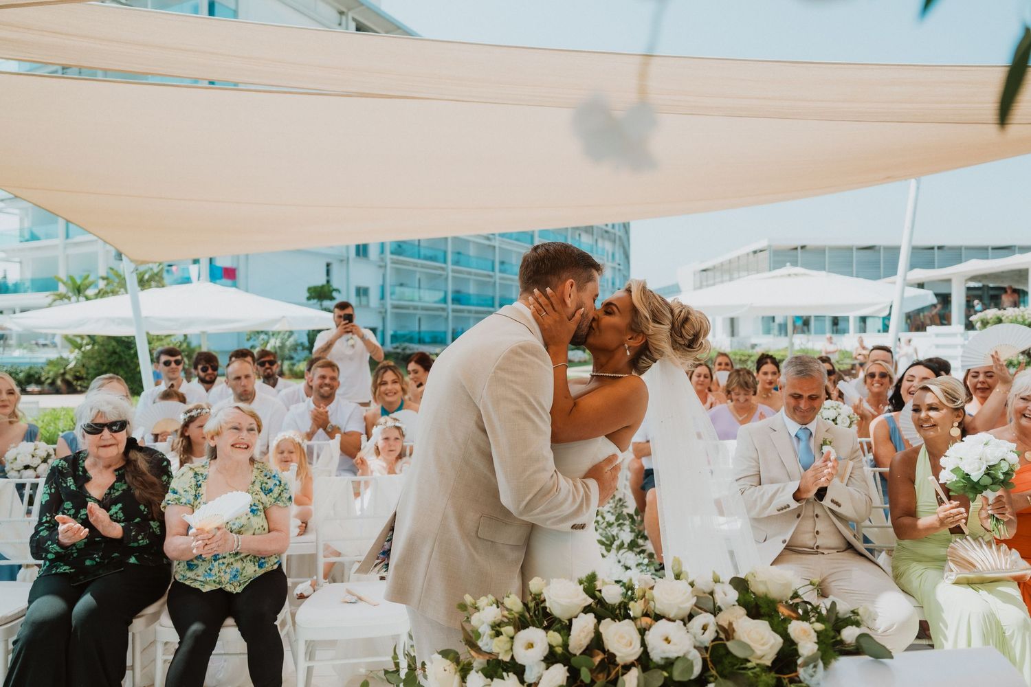 Bride and groom share first kiss at outdoor beach wedding ceremony with guests dressed in white applauding.