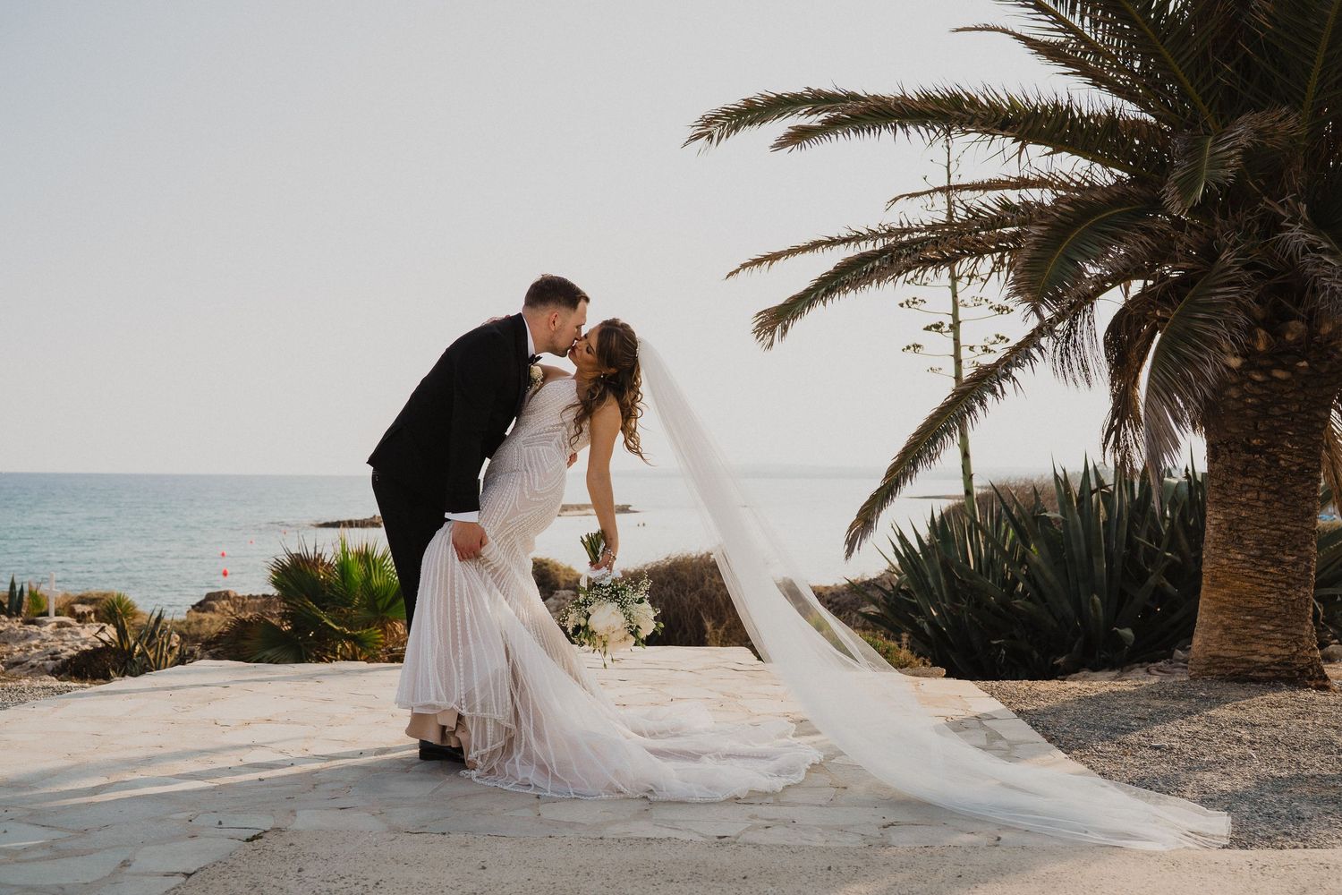 Groom dips bride for romantic kiss outdoors with ocean view and palm tree at coastal wedding venue.