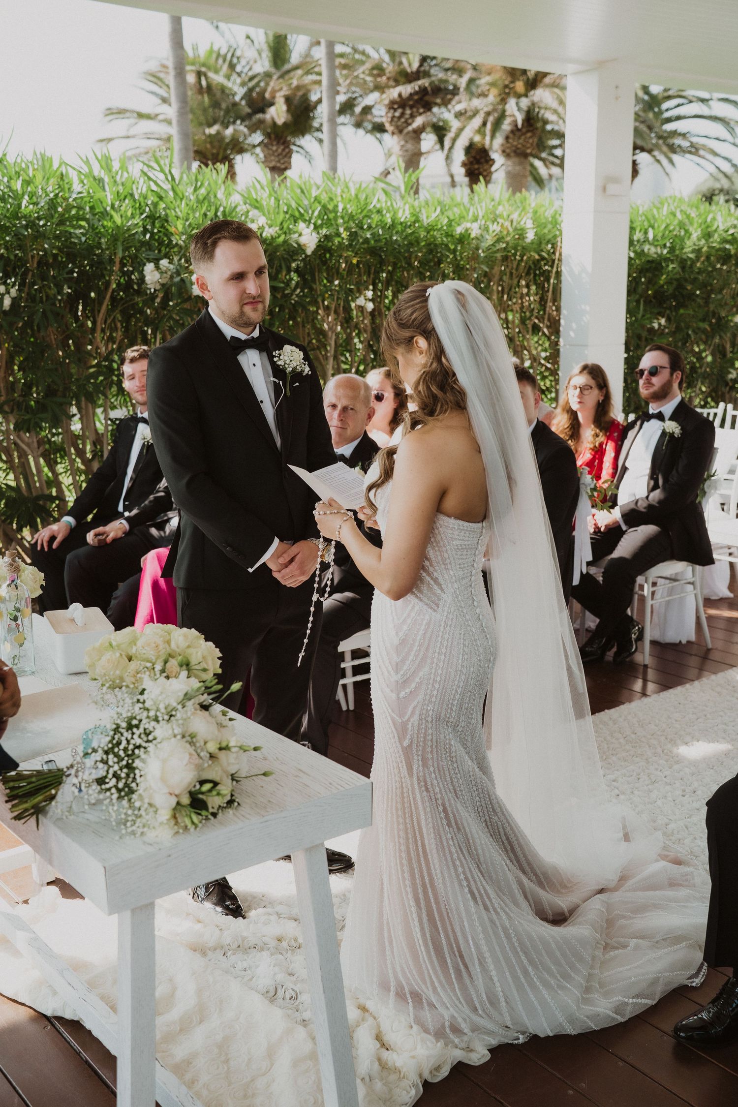 Couple exchanging vows at elegant outdoor wedding ceremony with lush green backdrop and guests.