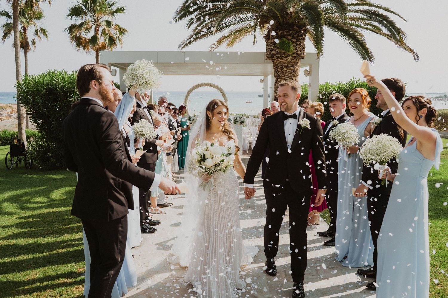 Bride and groom walk down the aisle as guests toss white confetti at an outdoor wedding ceremony.