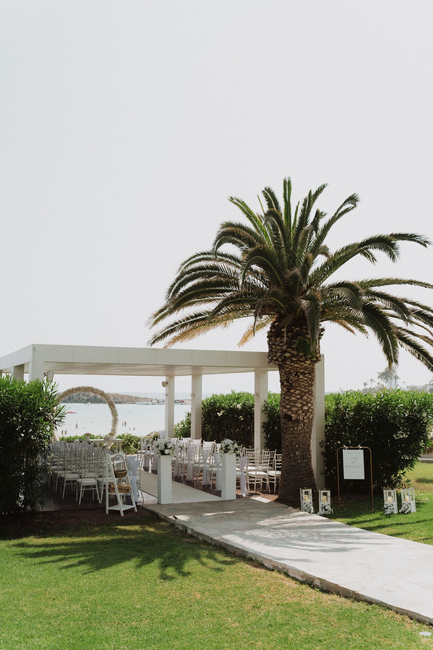 Outdoor wedding ceremony setup with white chairs, pergola, and palm tree at a garden venue.