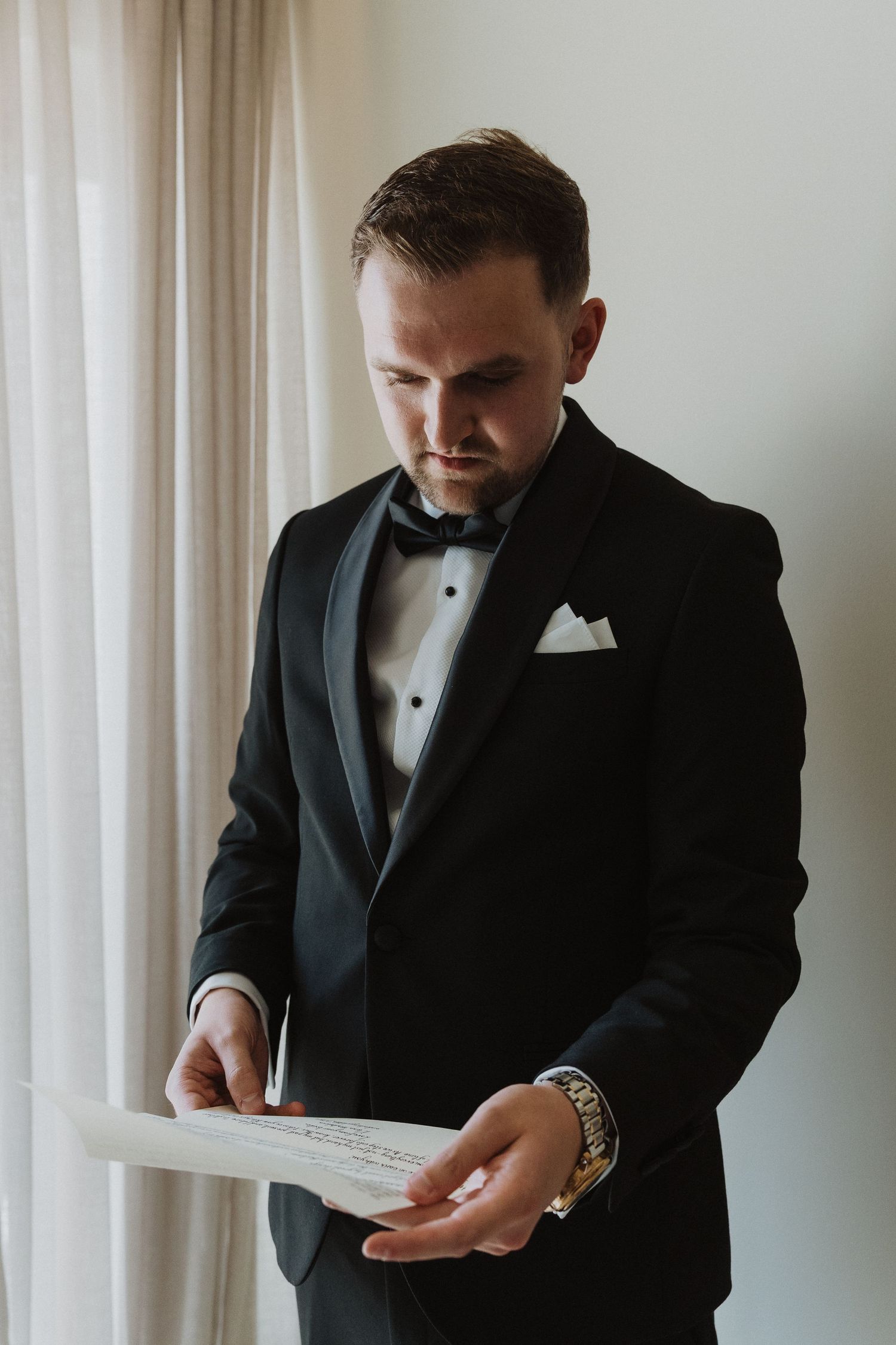 Groom in black tuxedo reading wedding vows on paper near window light.