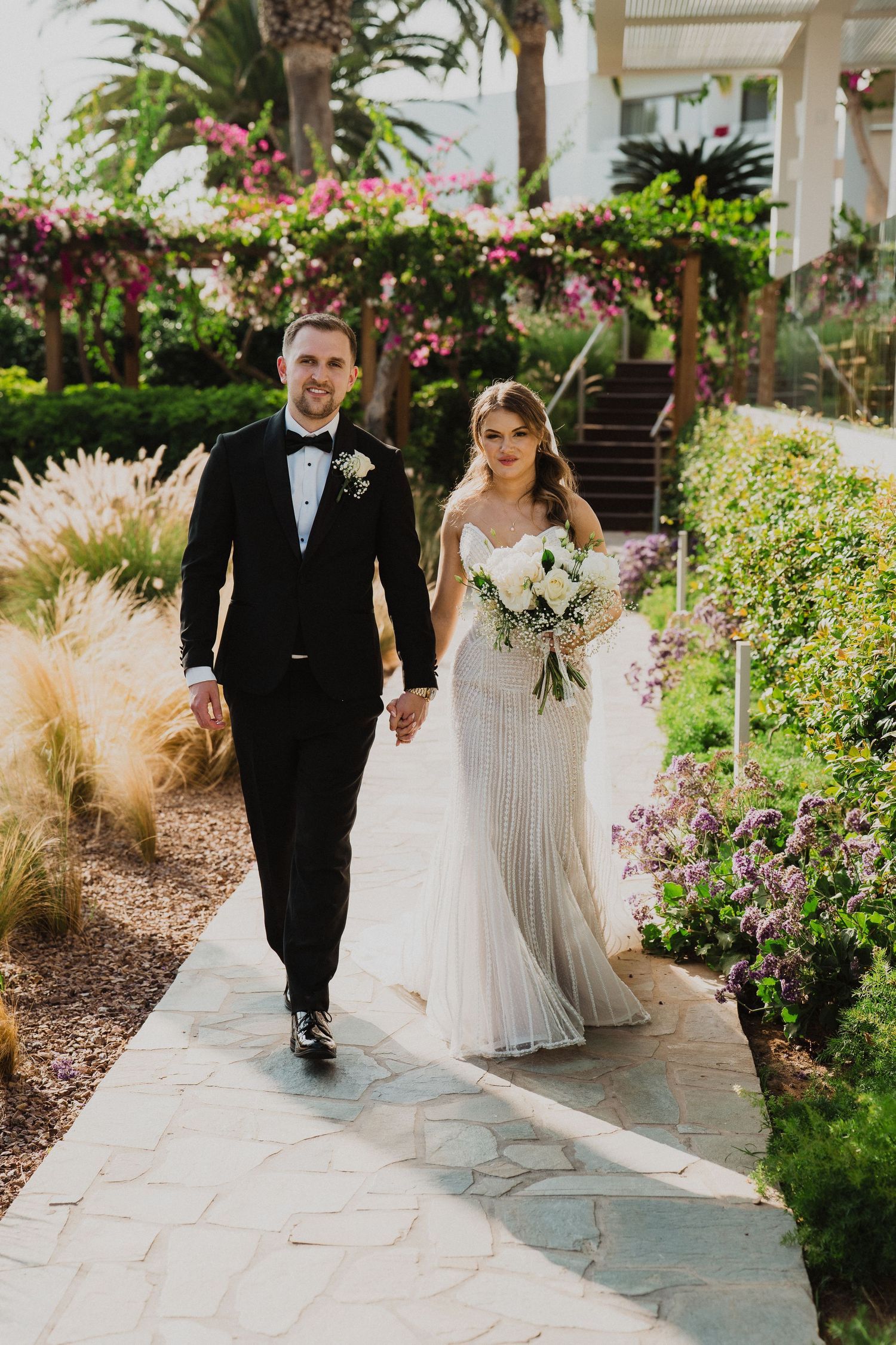 Smiling bride and groom walk hand-in-hand along a garden pathway surrounded by colorful flowers and lush greenery.