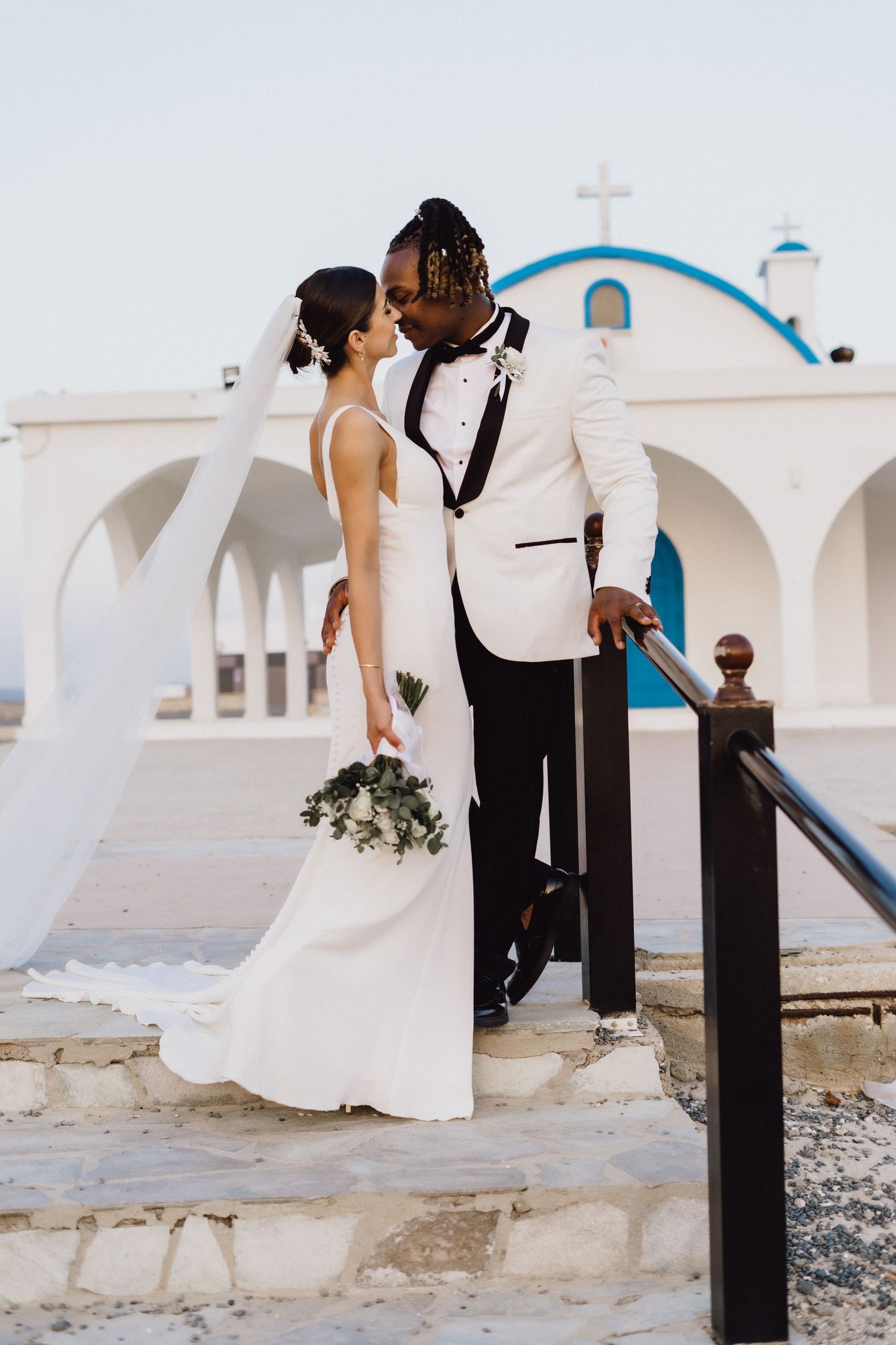 A romantic wedding couple embraces on white stone steps against a Mediterranean church with blue dome.