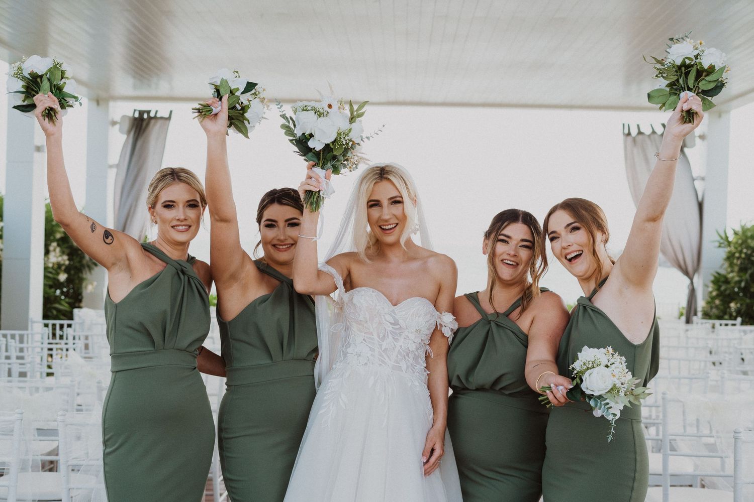 Bridesmaids in sage green dresses celebrate joyfully with raised bouquets.