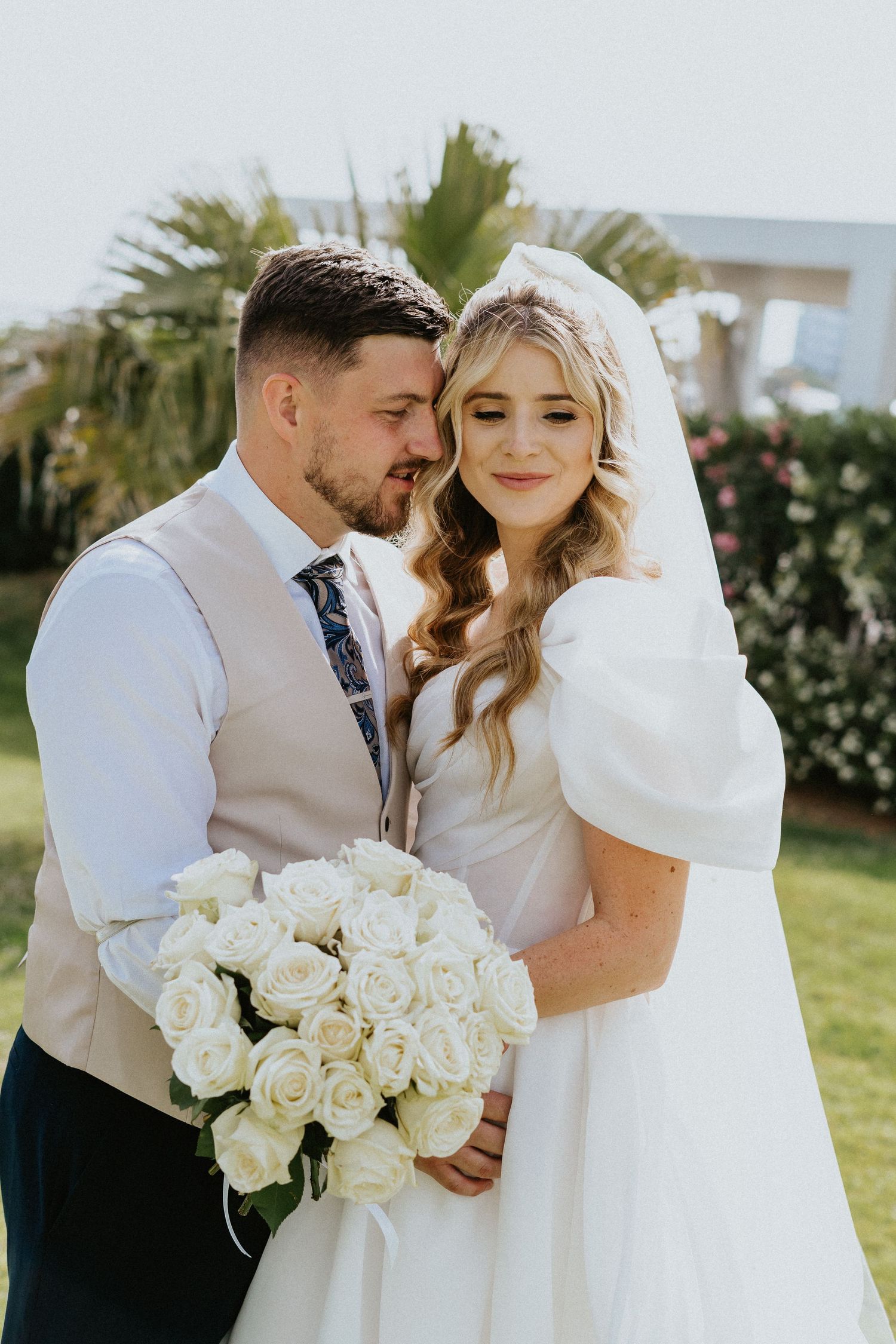A couple shares an intimate moment on their wedding day, with a beautiful bouquet of white roses in a sunny garden setting.