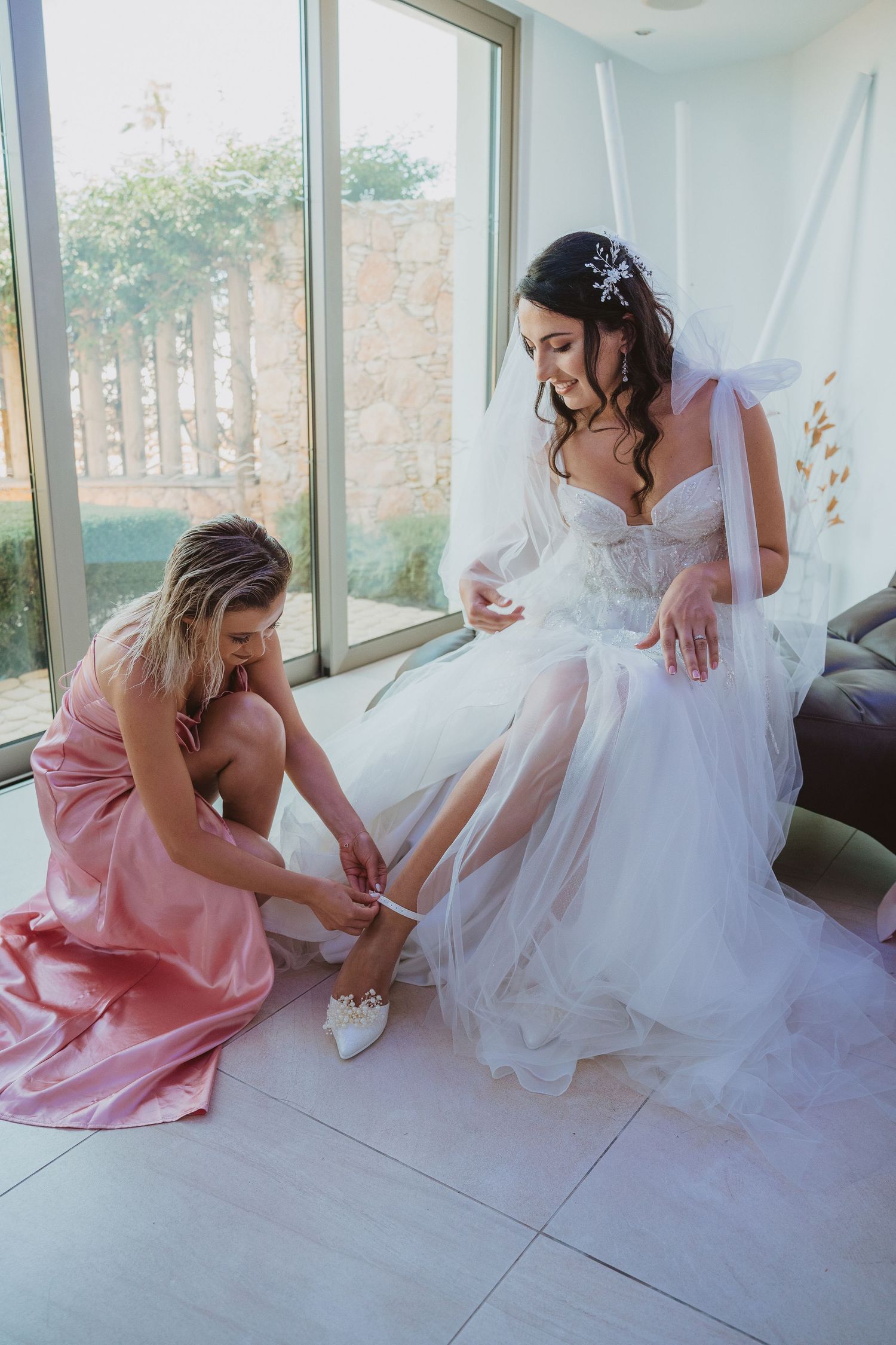Bridesmaid helps bride with her wedding shoes in modern bright getting-ready room.