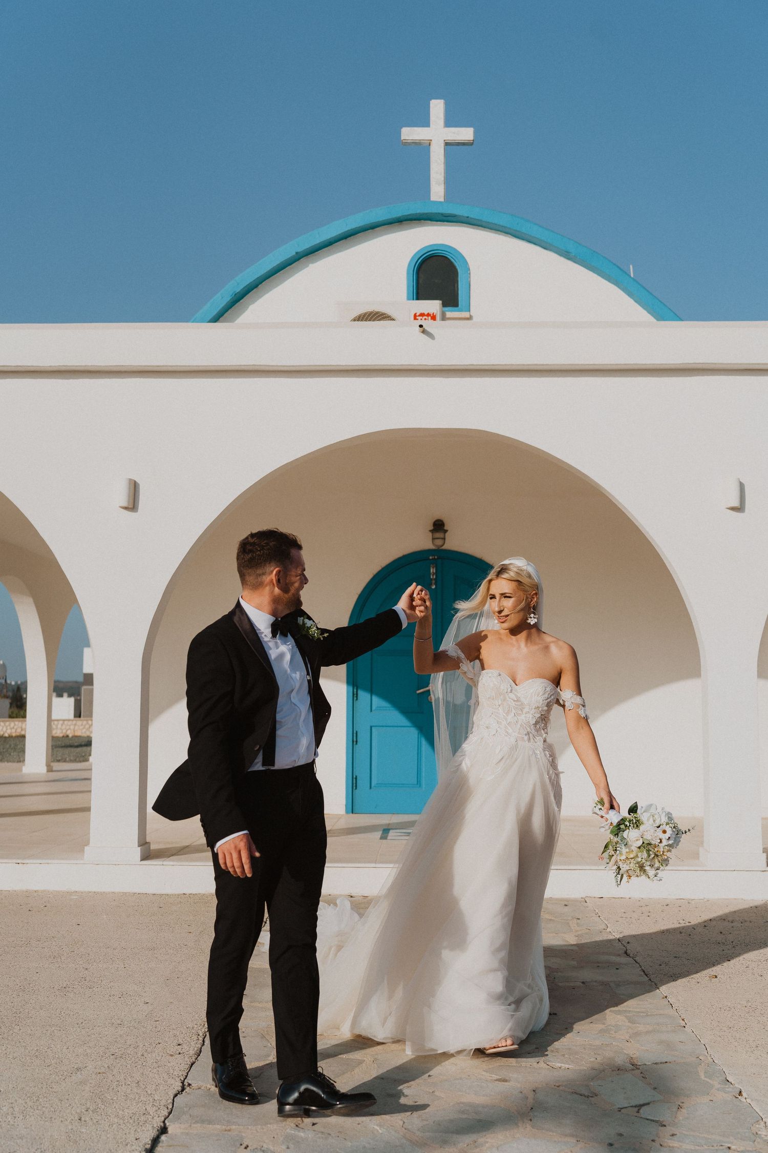Wedding couple poses at Mediterranean chapel with iconic blue door and white architecture.