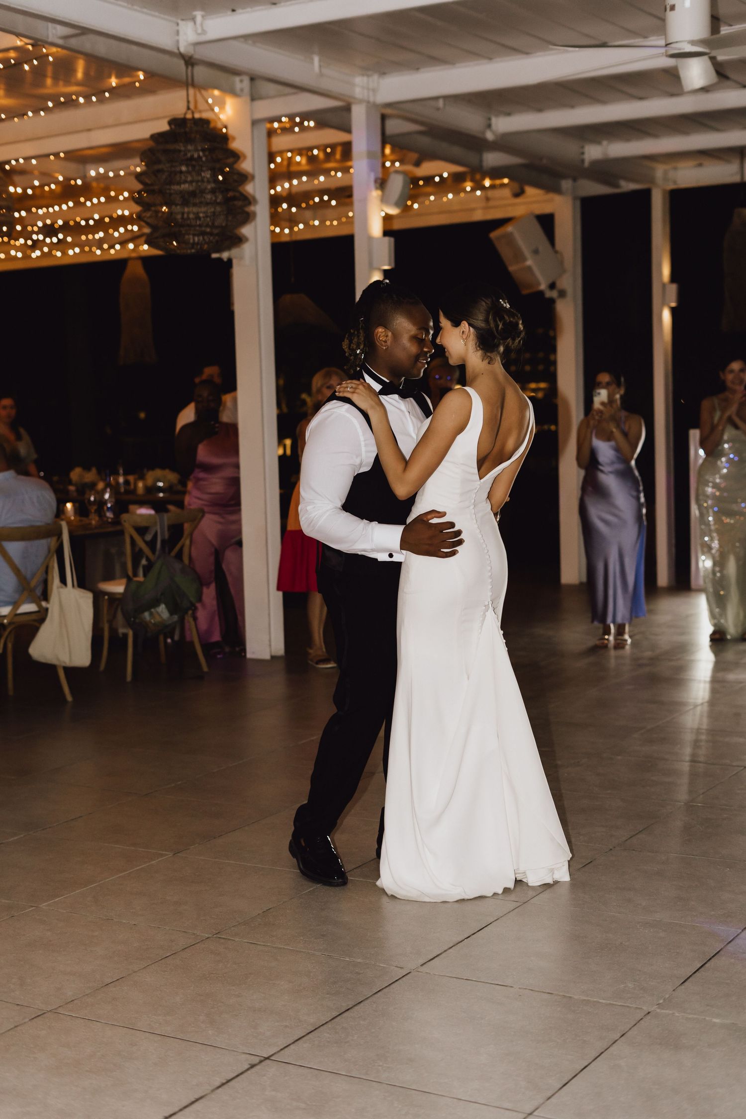 Couple shares their first dance on a tiled floor under romantic string lights at wedding reception.