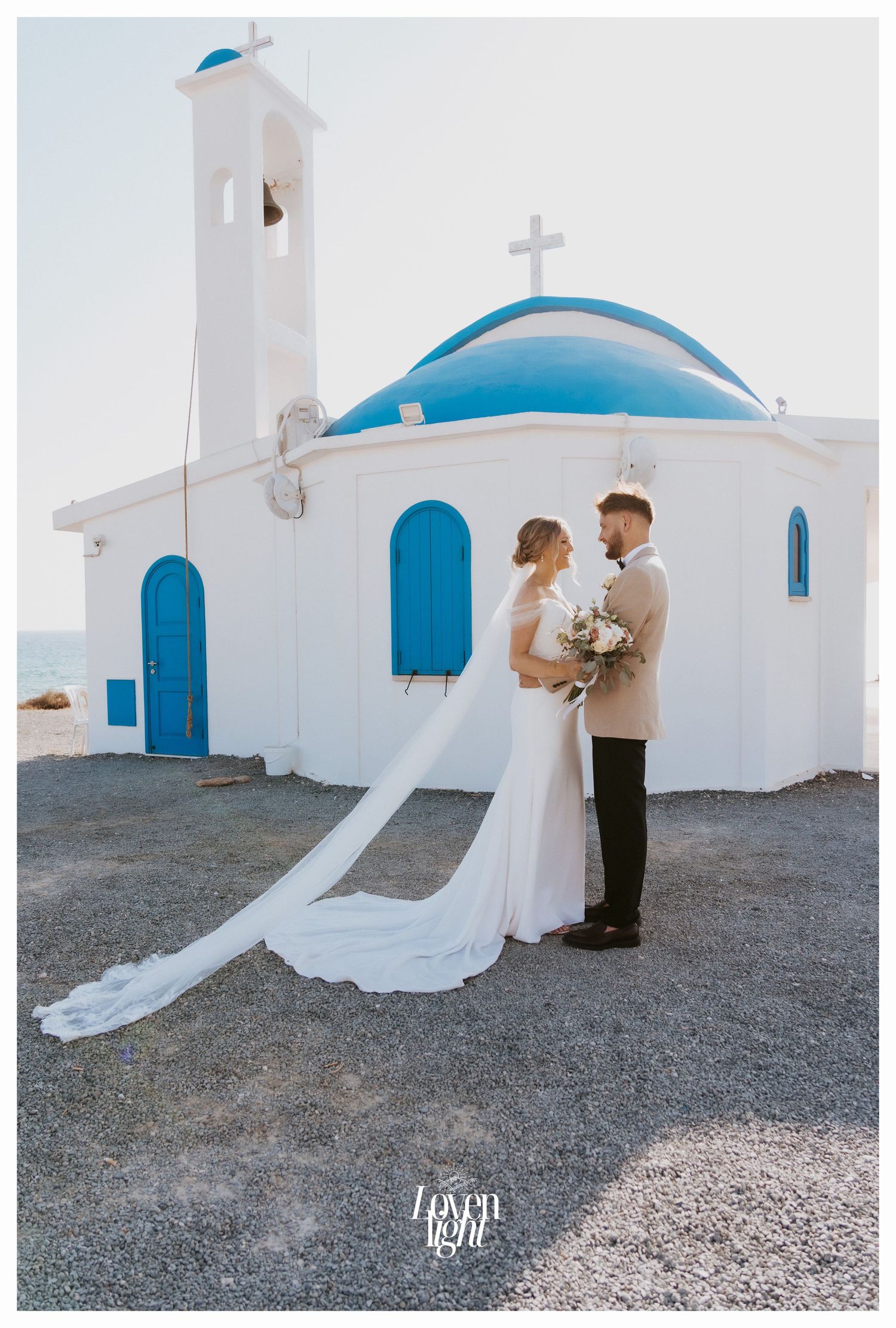 Wedding couple poses in front of a traditional white Greek church with blue dome and bell tower.